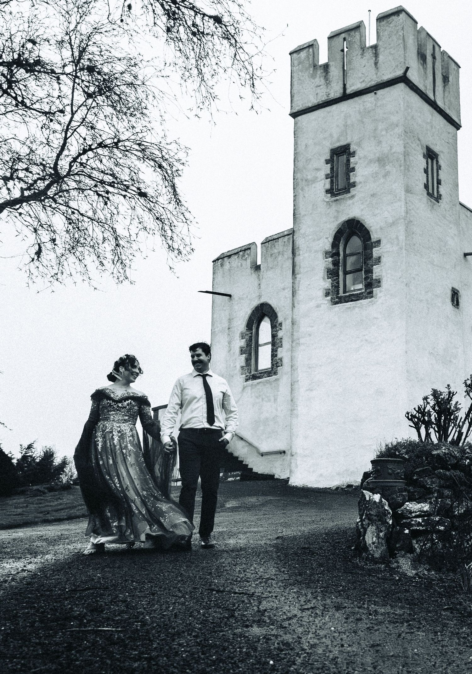 Bride in embellished gown and groom walk hand in hand beside historic white castle tower.