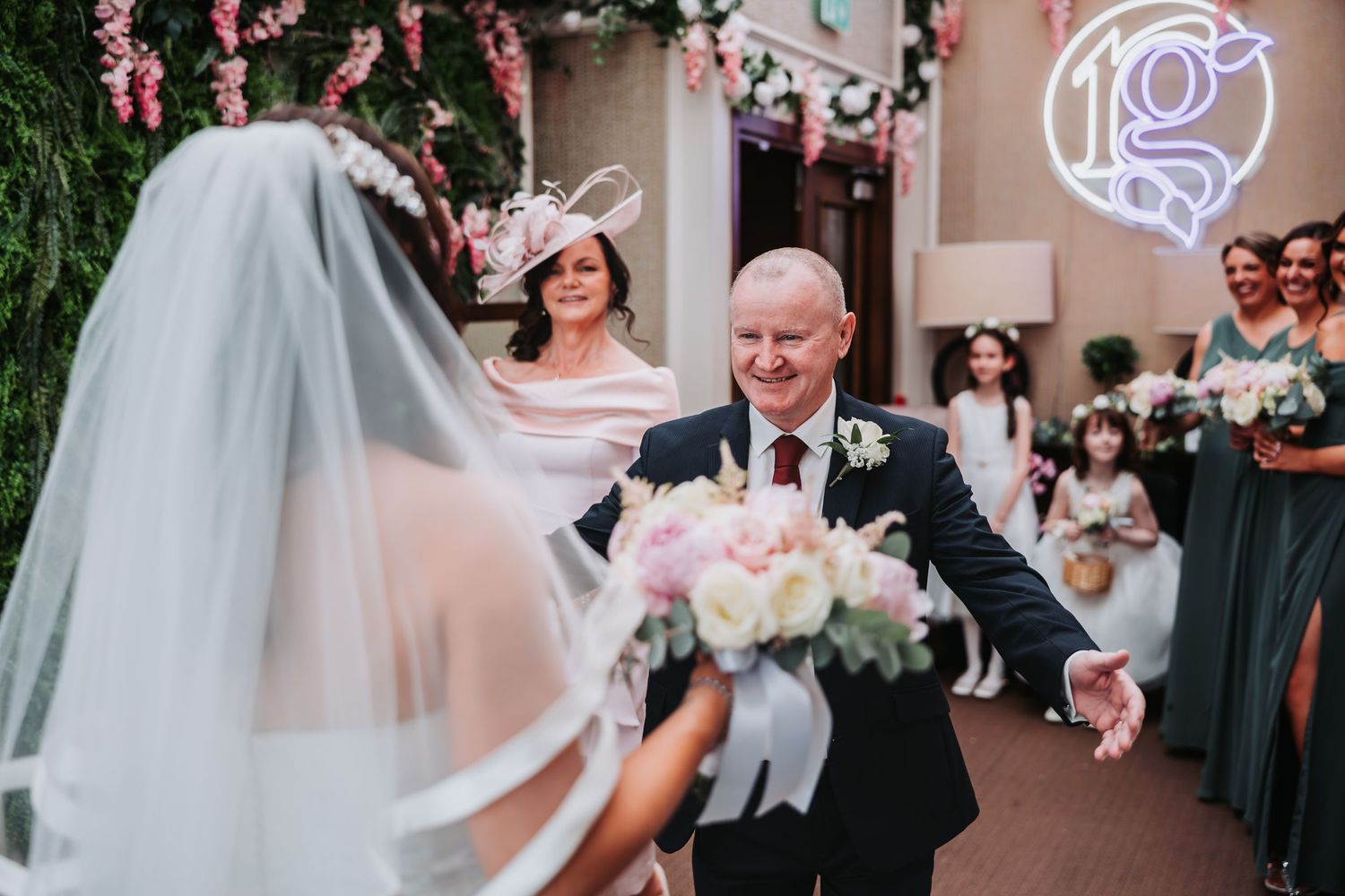 Father of the bride smiling and reaching for his daughter's hand at a floral wedding ceremony aisle.
