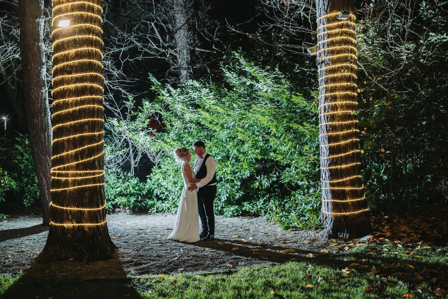 A couple shares an intimate moment between two trees wrapped in twinkling lights at night.