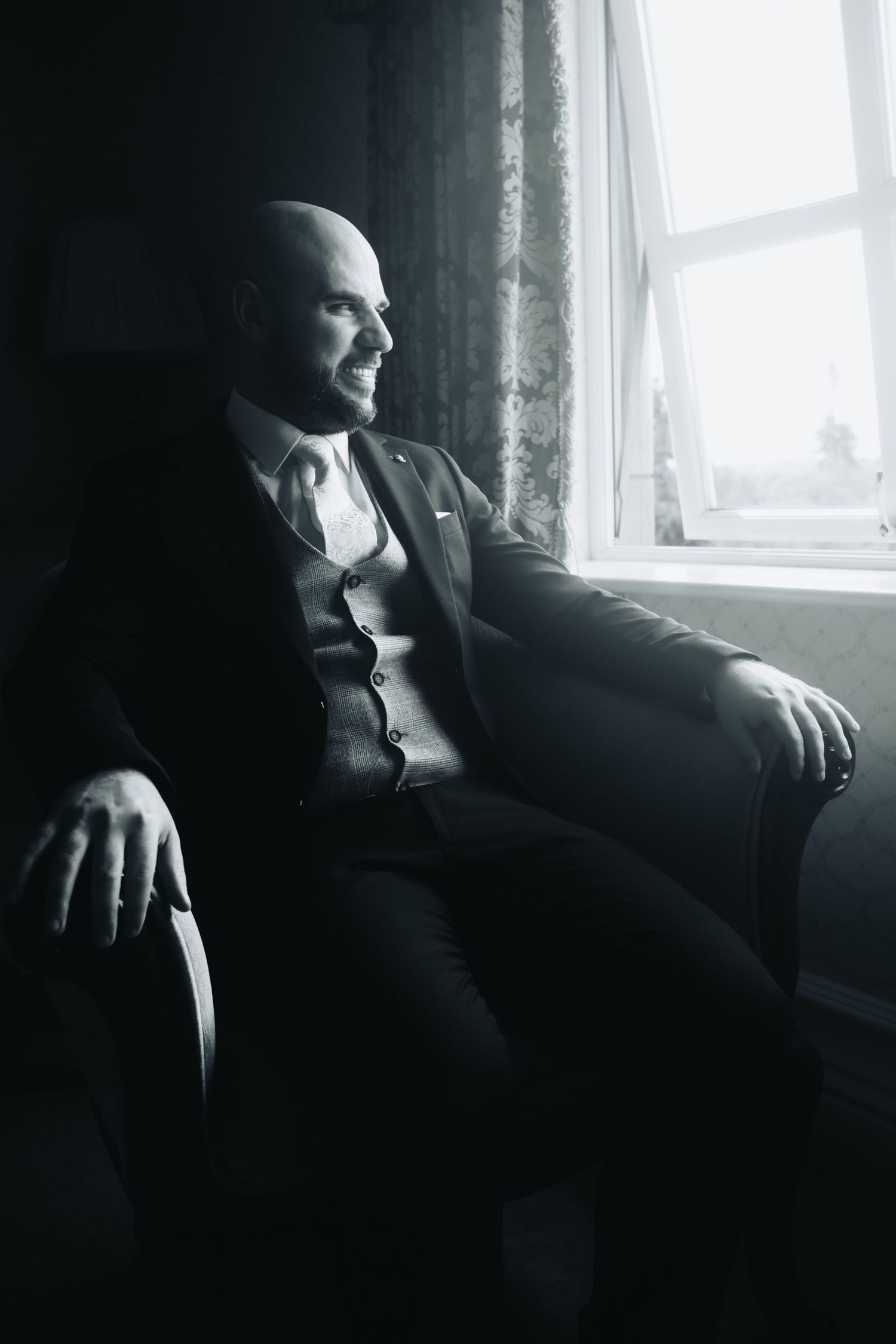 Bald groom in a three-piece suit sits in an armchair by a window, smiling in a black and white portrait.