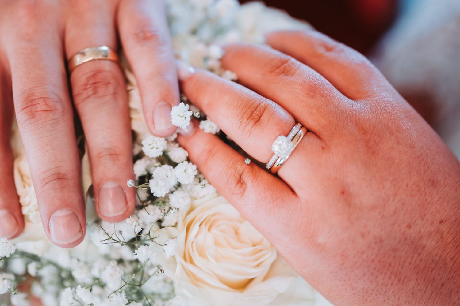 Close-up of newlyweds hands showing wedding bands and diamond engagement ring over white roses.