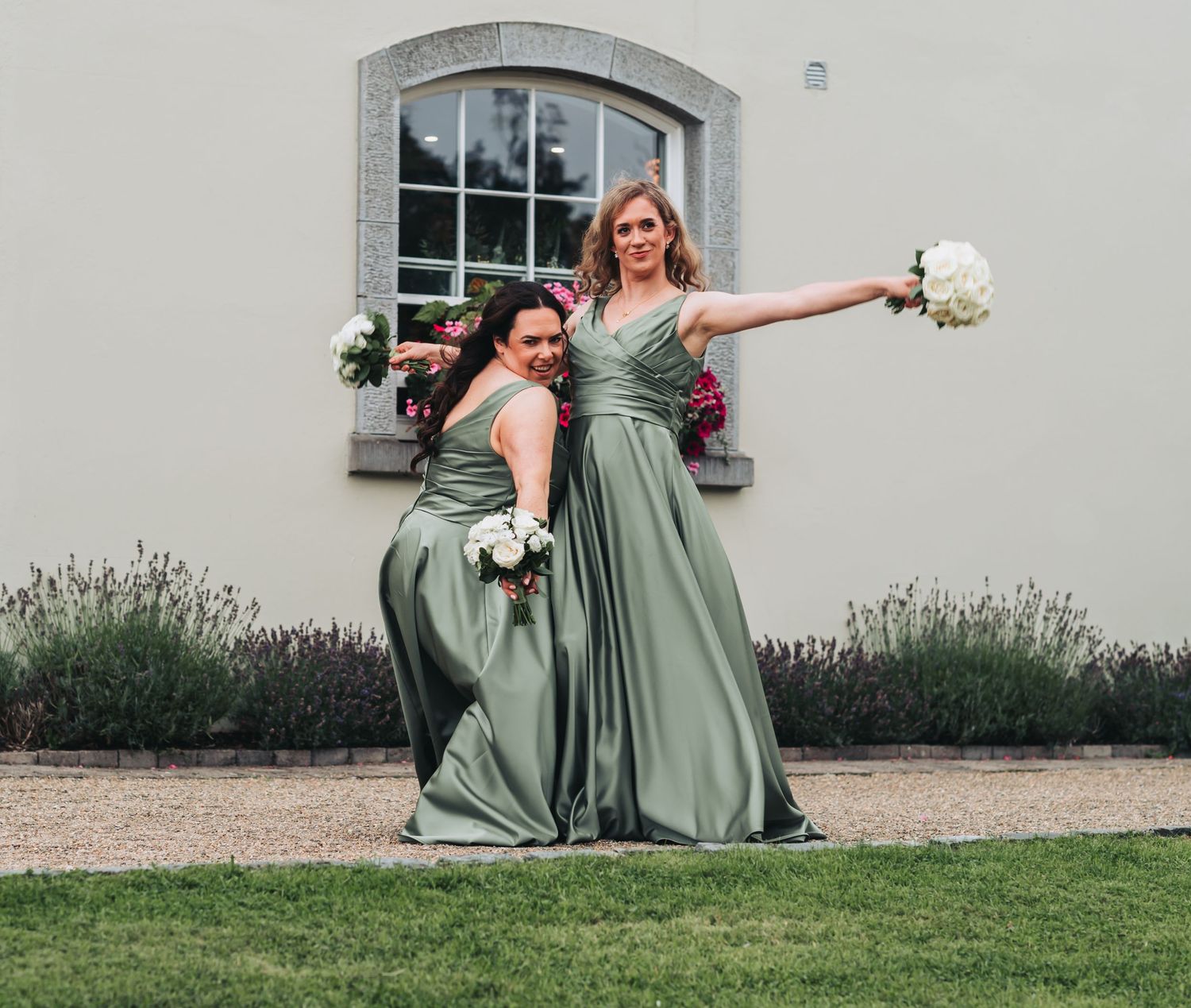 Two bridesmaids in sage green dresses posing playfully with white bouquets outside a country wedding venue.