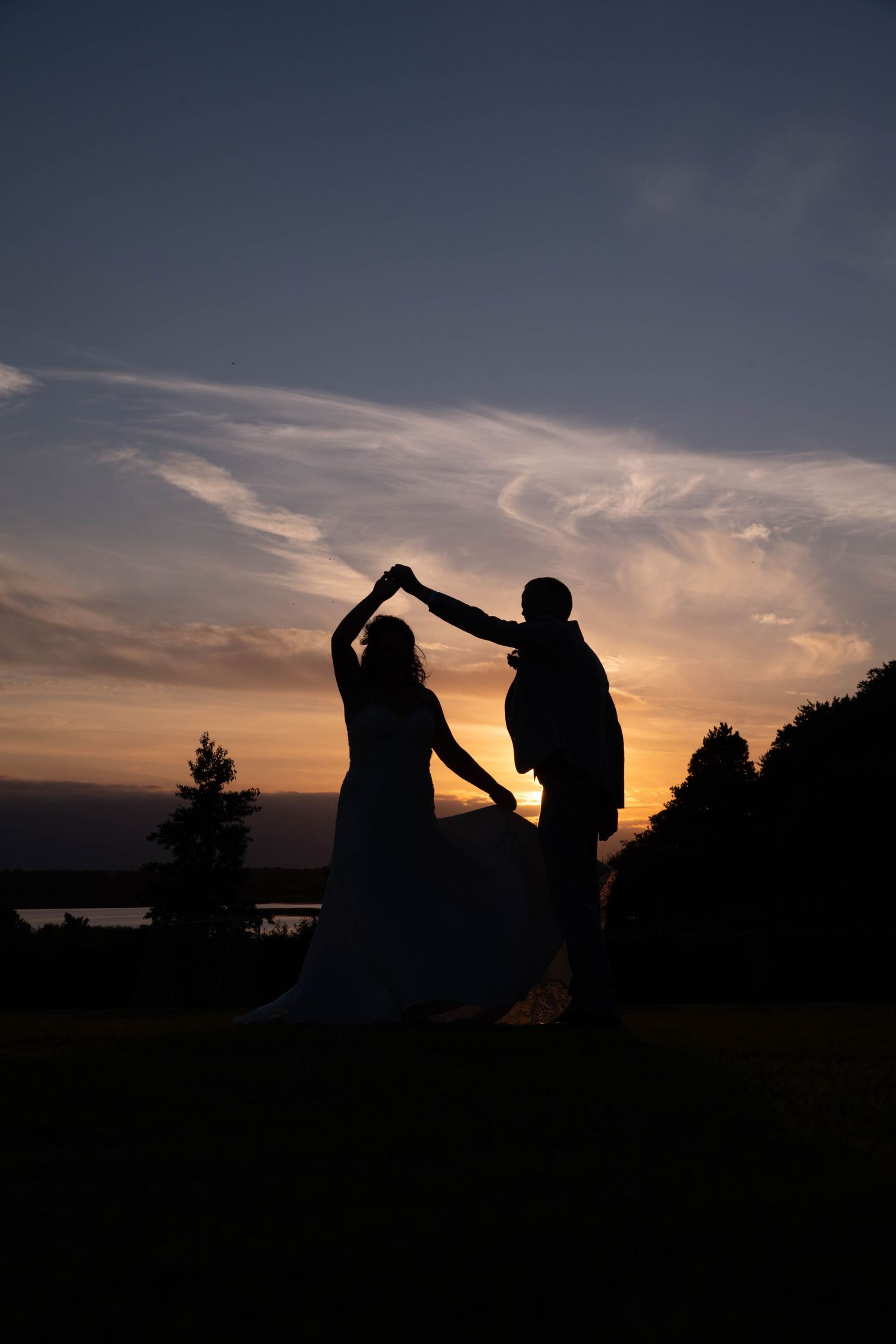 Silhouette of a bride and groom dancing at sunset with vibrant orange sky and trees in the background.