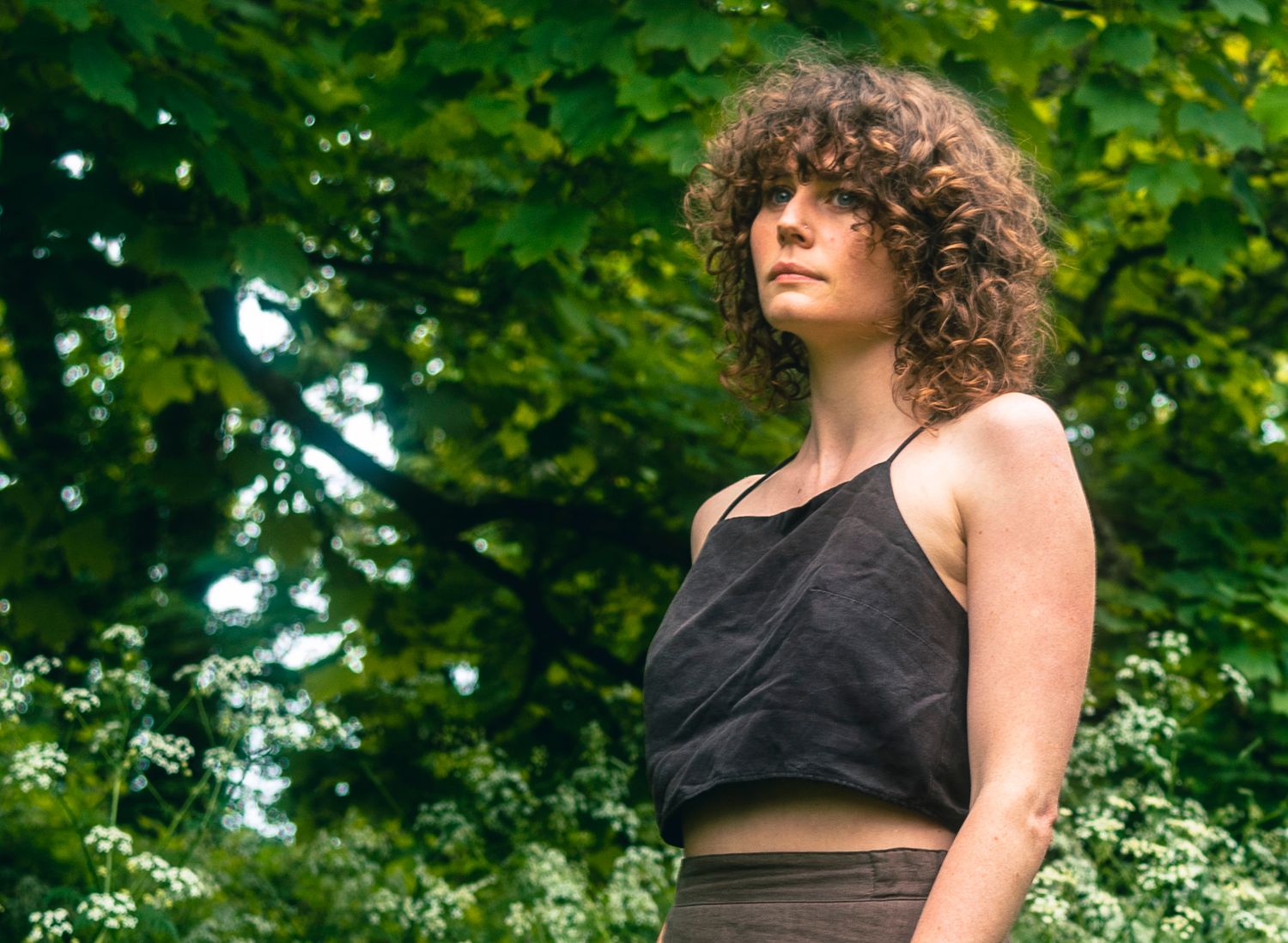Young woman with curly hair in black crop top looking up among lush green trees outdoors.
