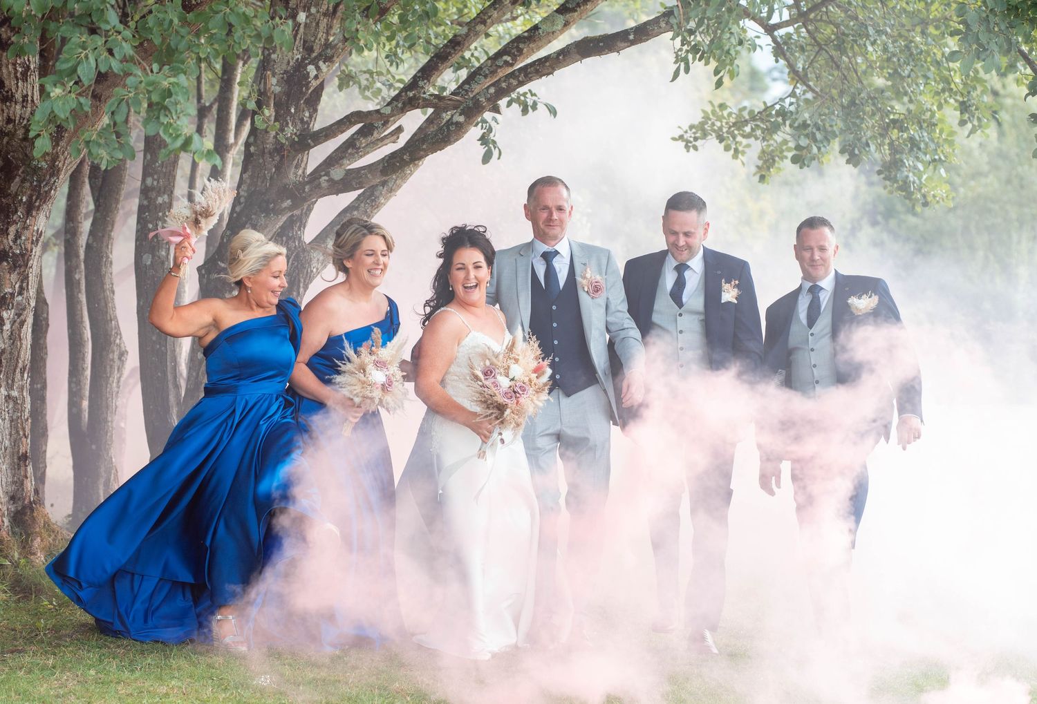 Wedding party celebration with pink smoke effects among trees outdoors.