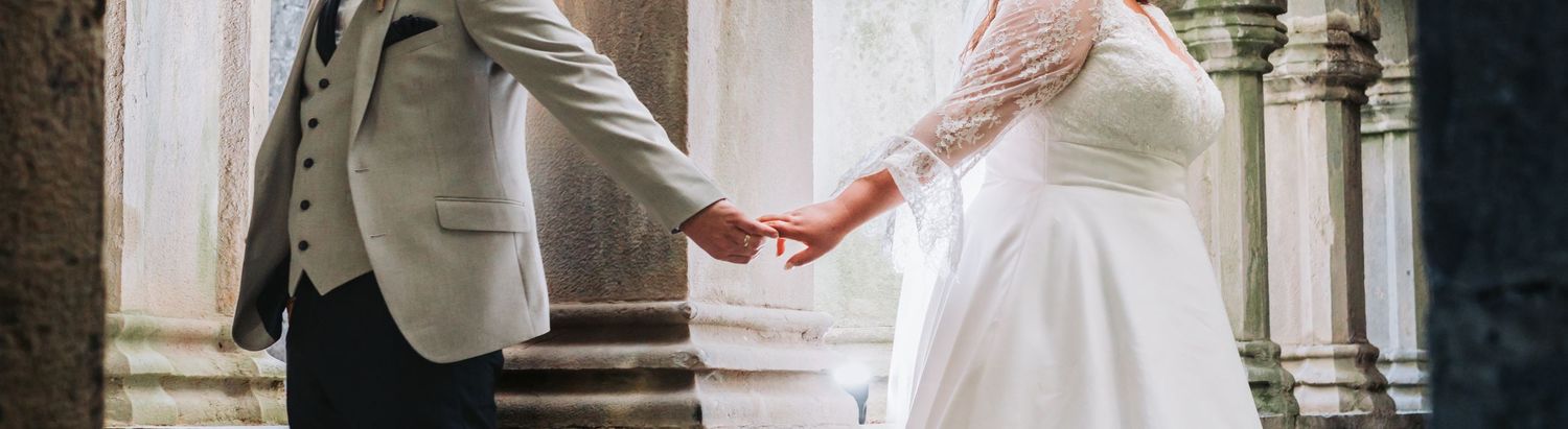 Bride and groom hold hands in elegant venue, bride in lace dress and groom in grey suit.