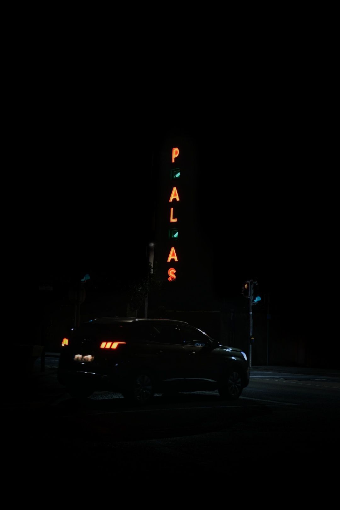 Night scene with a car parked near a glowing PALAS neon sign on a dark street.