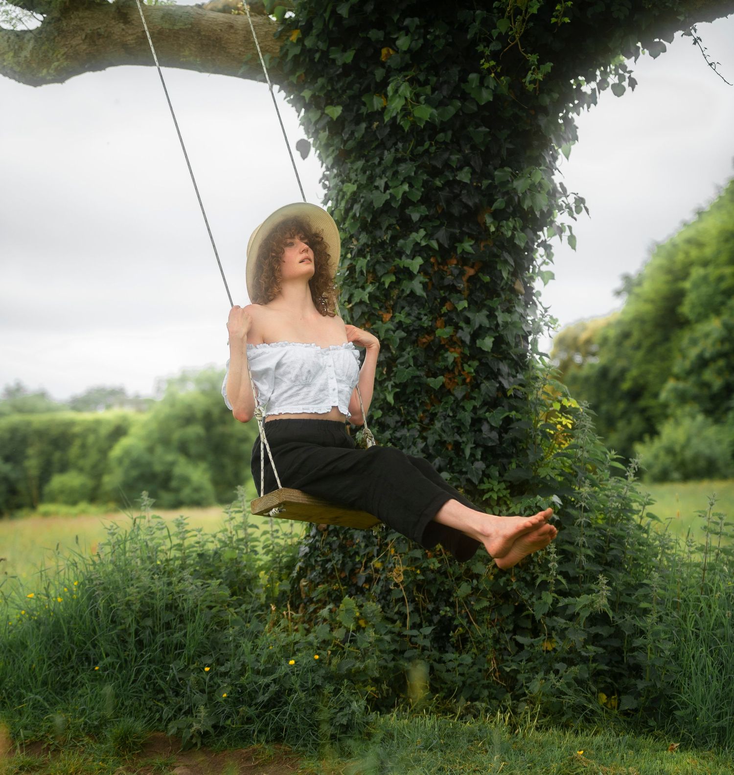 Young woman in sun hat sitting on tree swing surrounded by lush green ivy and garden.
