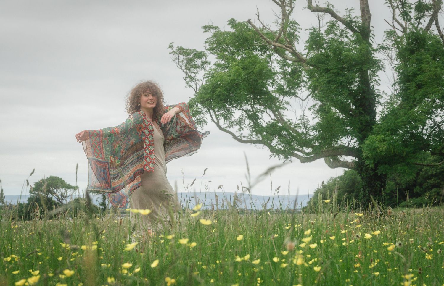 Woman in colorful kimono wrap stands in wildflower meadow with yellow blooms and windswept tree under cloudy sky.
