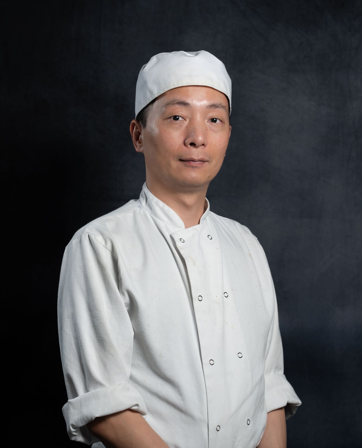 A professional chef in white uniform and cap poses for a portrait against a dark background.