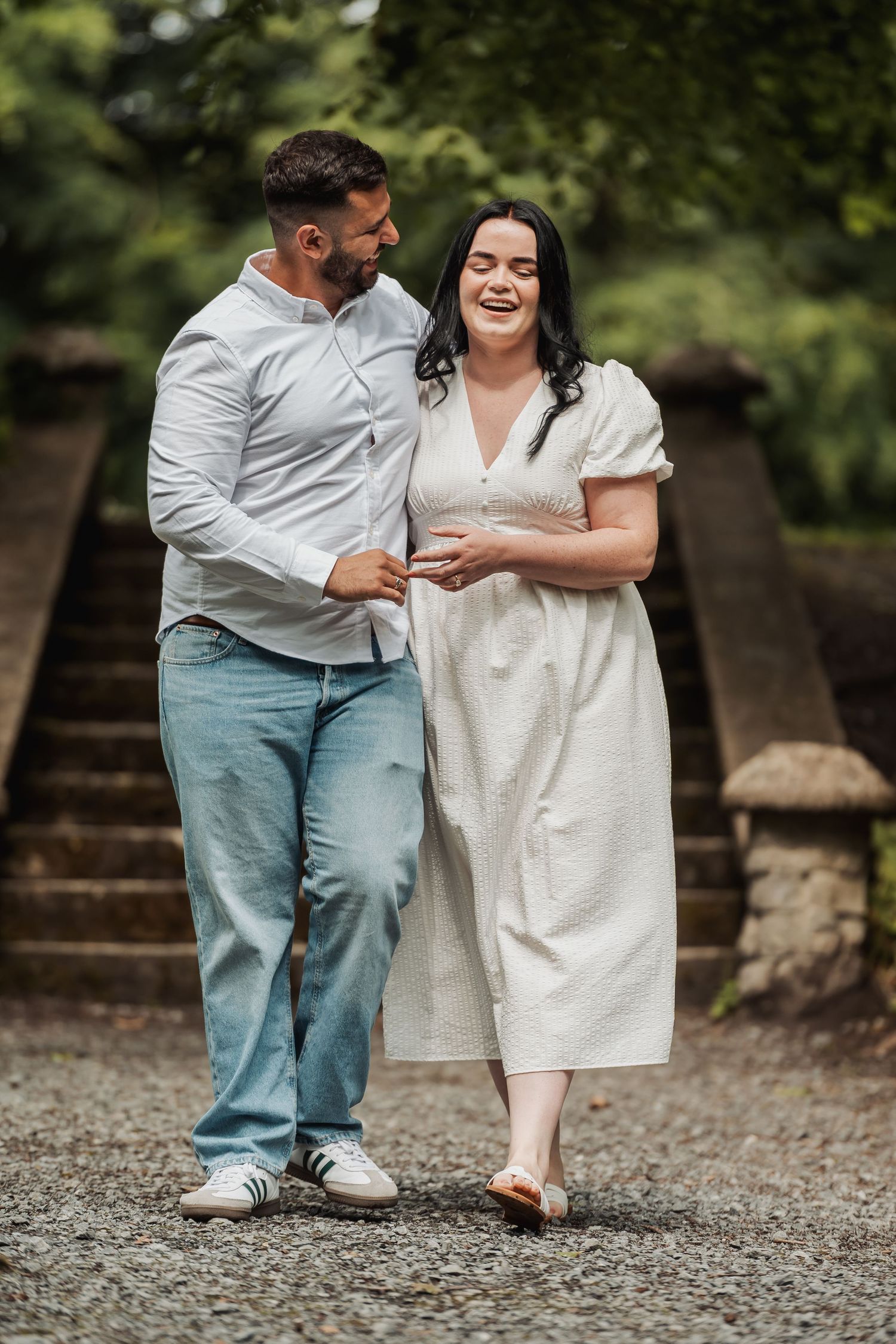 A happy couple walks together in a lush green park, the woman in a white dress laughing joyfully.