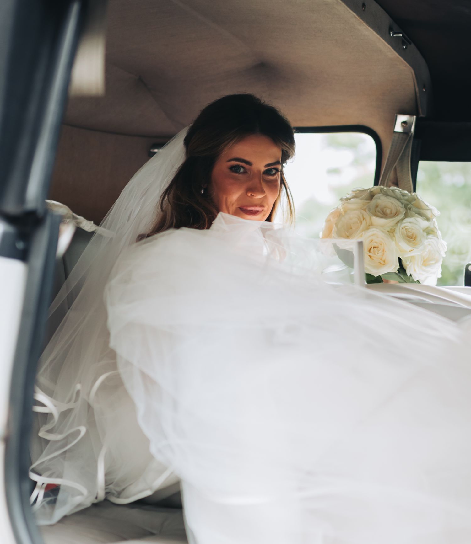 Radiant bride in white gown and veil holding bouquet inside vintage wedding car.