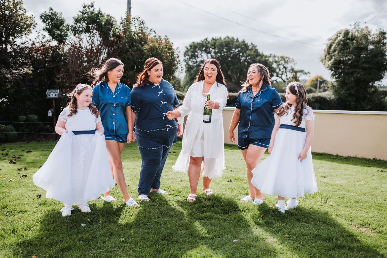 Bridesmaids in navy robes and flower girls in white dresses celebrate outdoors with champagne.