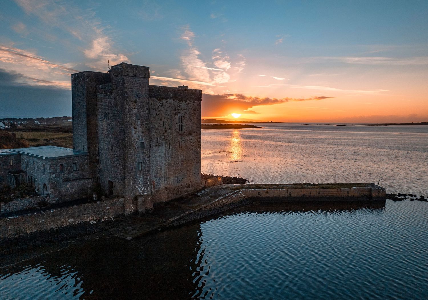 Aerial view of a medieval stone castle at sunset beside calm waters with vibrant orange sky reflections.