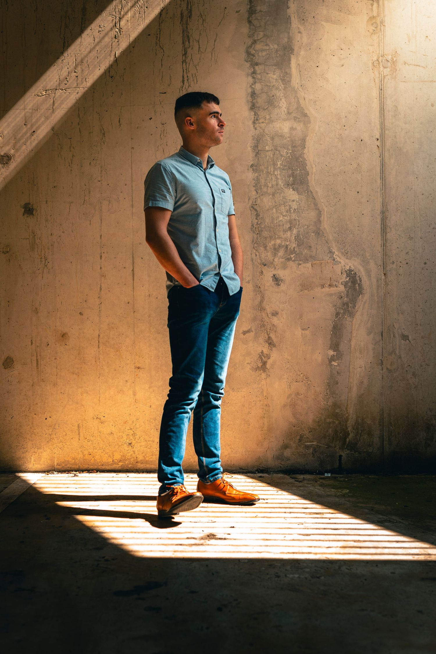 Young man in blue shirt and jeans standing in sunlit concrete stairwell.