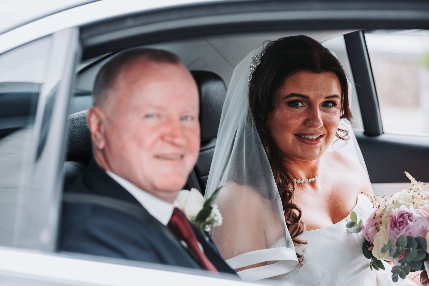 Bride in white gown and veil holding bouquet sits beside older man in suit inside a car on wedding day.