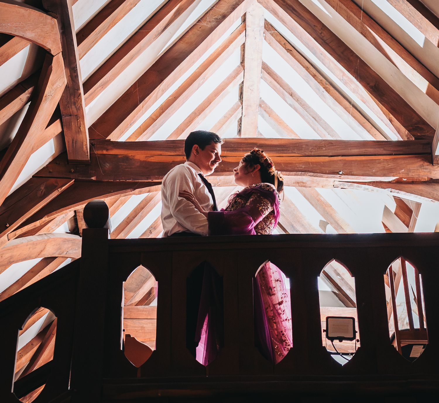 Couple sharing a romantic moment on a wooden balcony beneath rustic timber roof beams in warm light.
