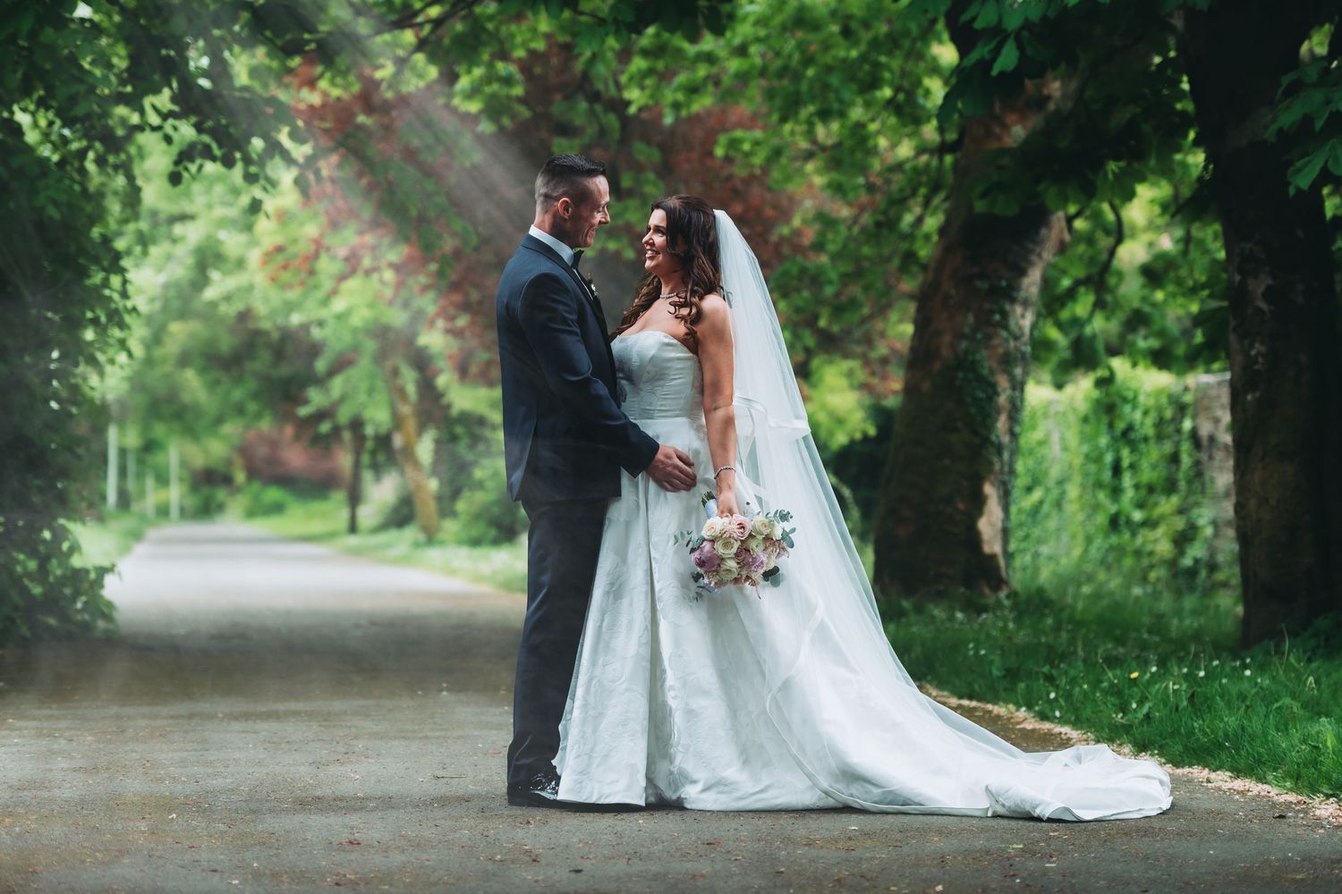 Bride and groom share a romantic moment on a tree-lined path, with soft sunlight rays filtering through lush green foliage.