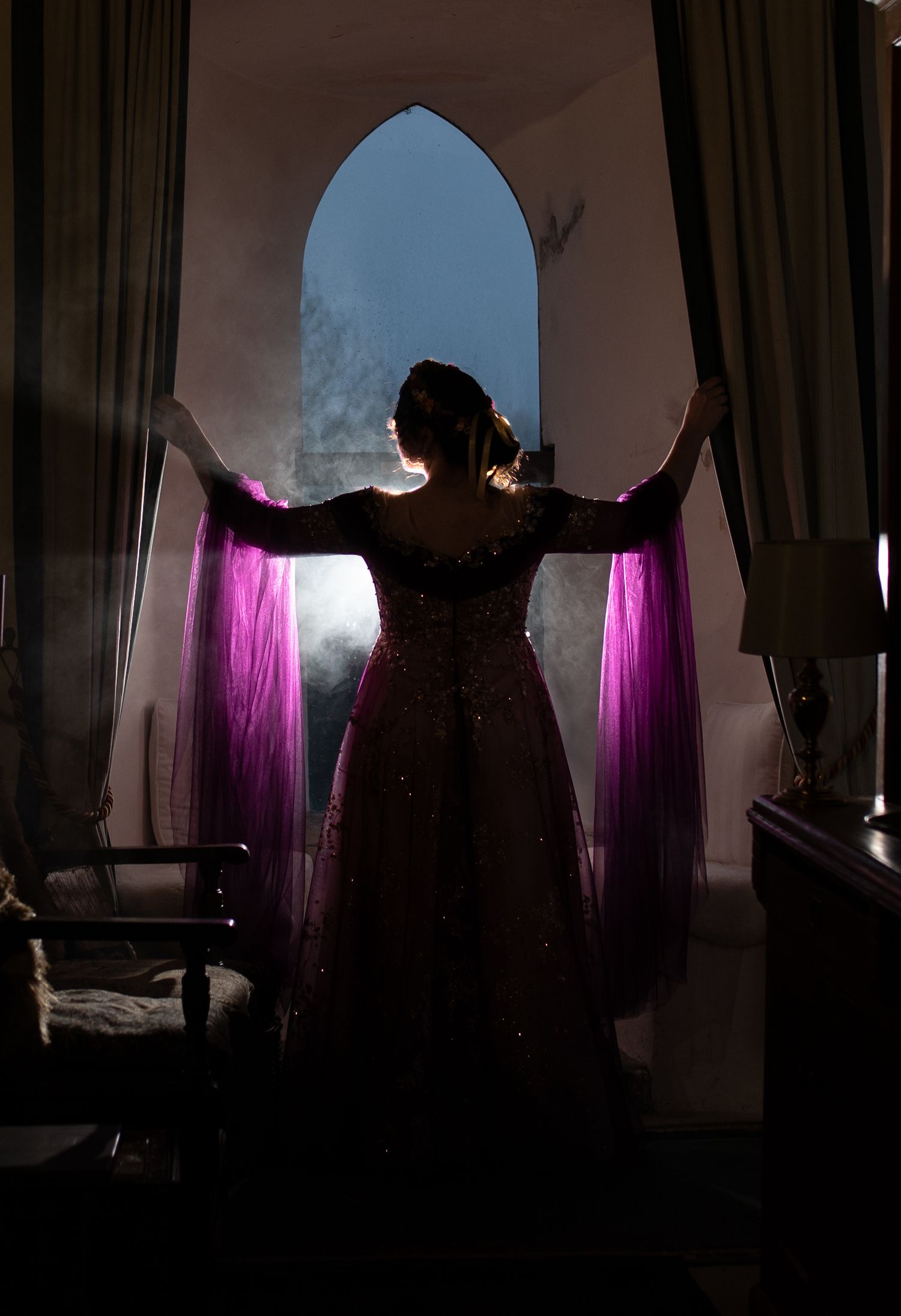 Woman in elegant pink gown with purple dupatta stands silhouetted before an arched window in dim room.