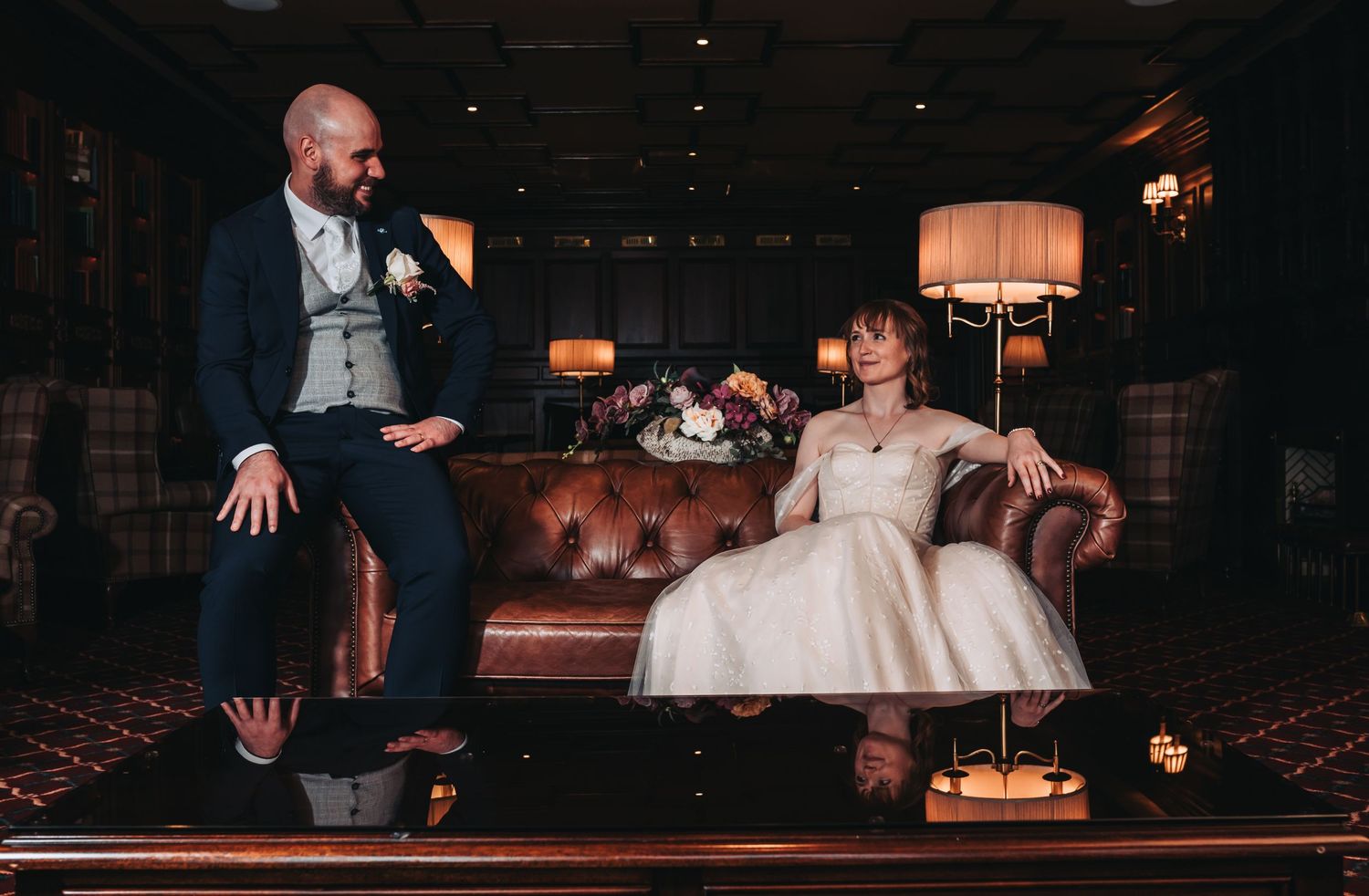 Bride and groom share a relaxed moment seated on a leather sofa in a warmly lit wedding venue.