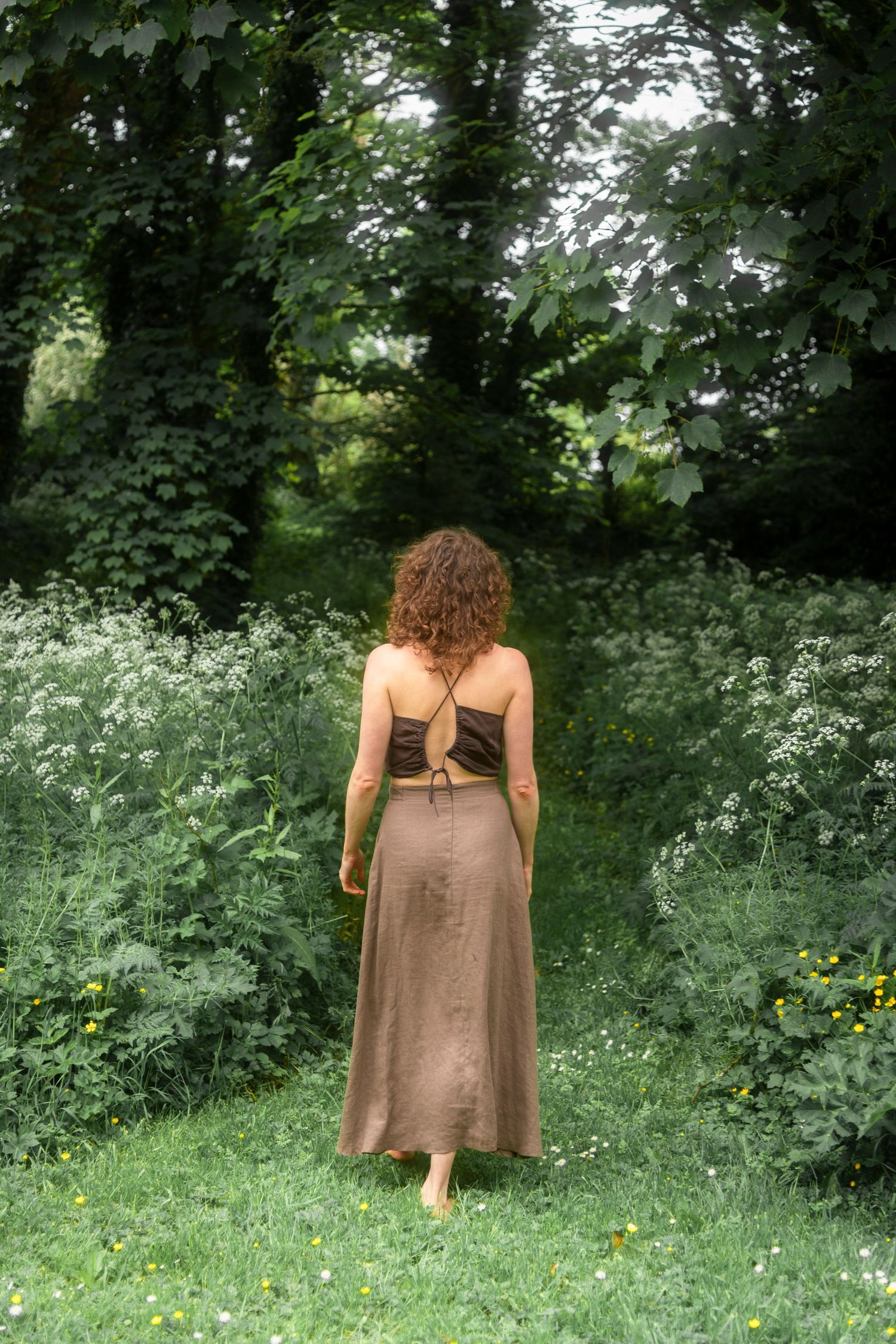 Woman in brown slip dress and crop top walking barefoot into lush forest.
