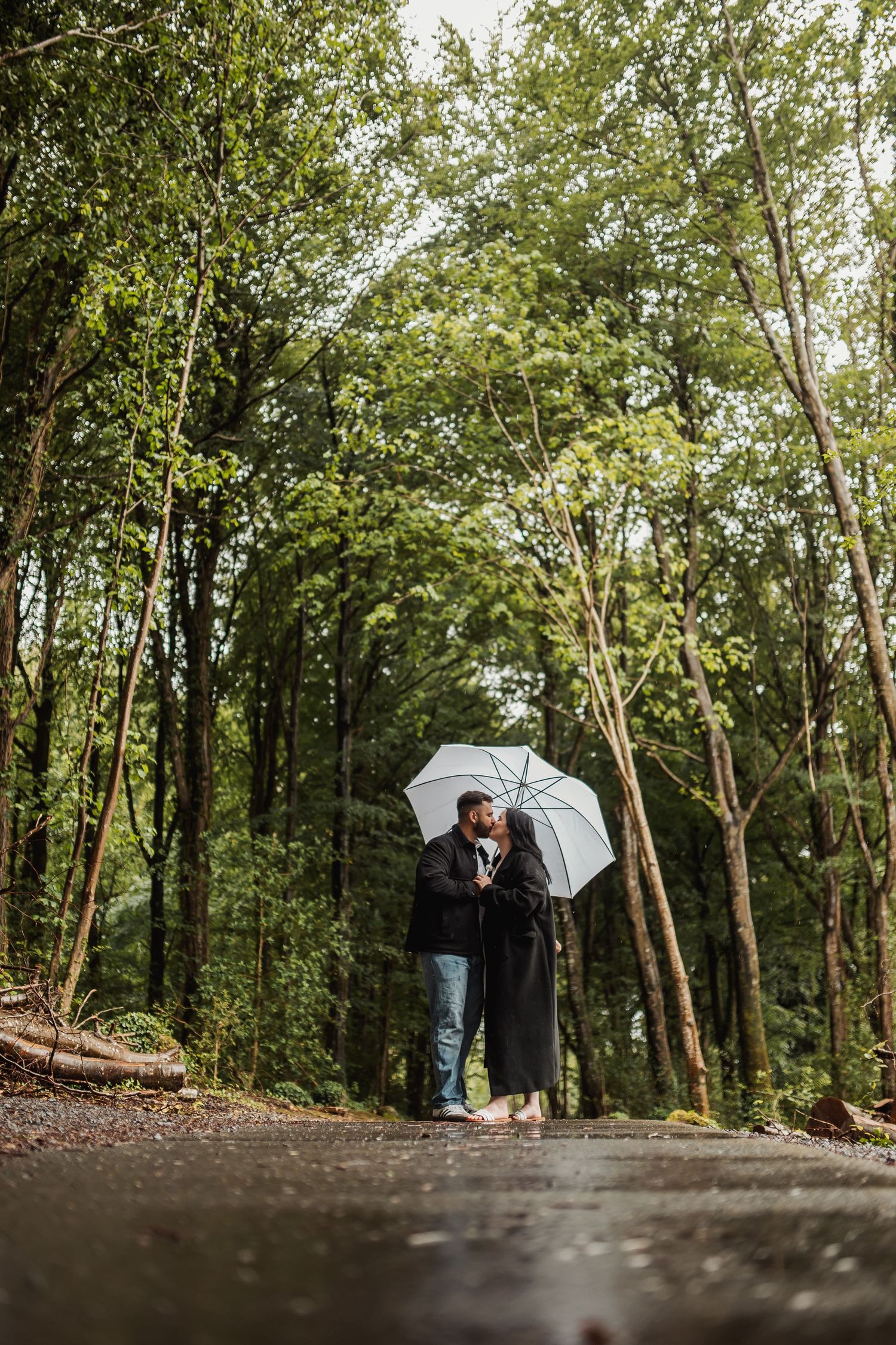 Couple sharing a romantic kiss under a white umbrella on a rainy forest path surrounded by lush green trees.