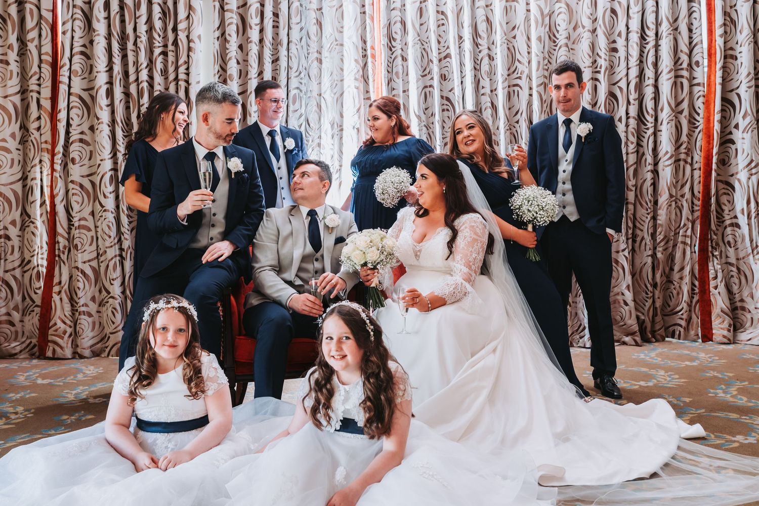 Wedding party poses with bride and groom seated, flanked by bridesmaids, groomsmen, and flower girls.