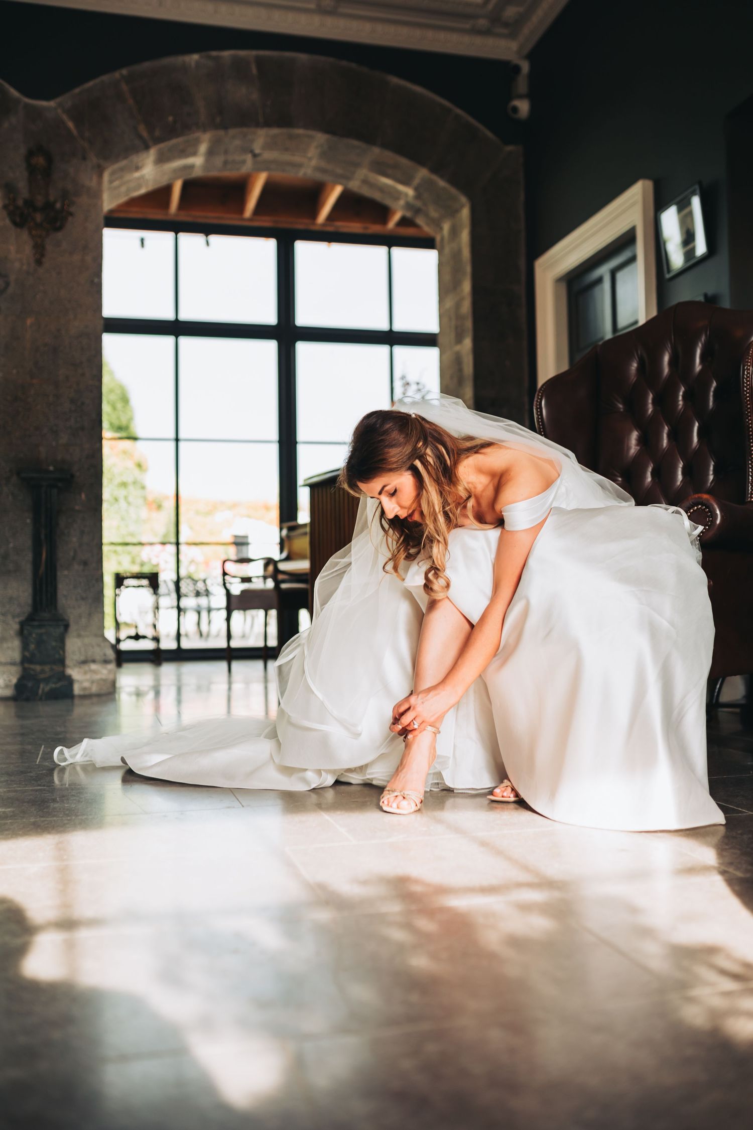 Bride in off-shoulder wedding gown adjusting shoes on floor in sunlit rustic venue interior.