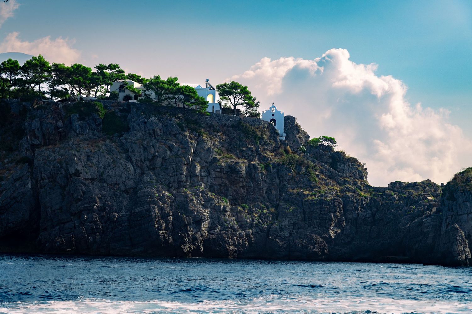 White chapel perched atop dramatic rocky coastal cliffs surrounded by pine trees and blue sea.