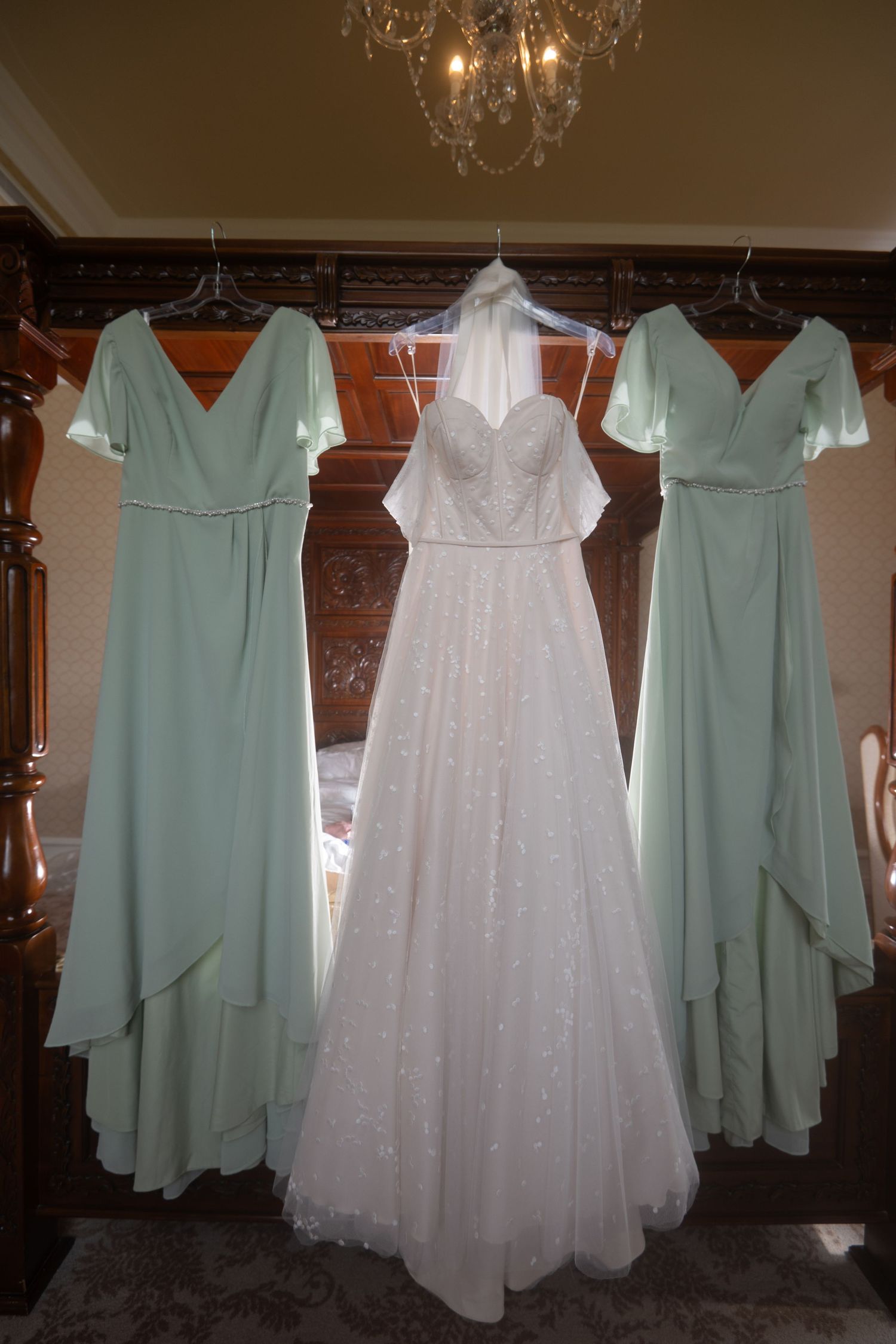 White wedding gown flanked by two sage green bridesmaid dresses hanging in an elegant room with chandelier.