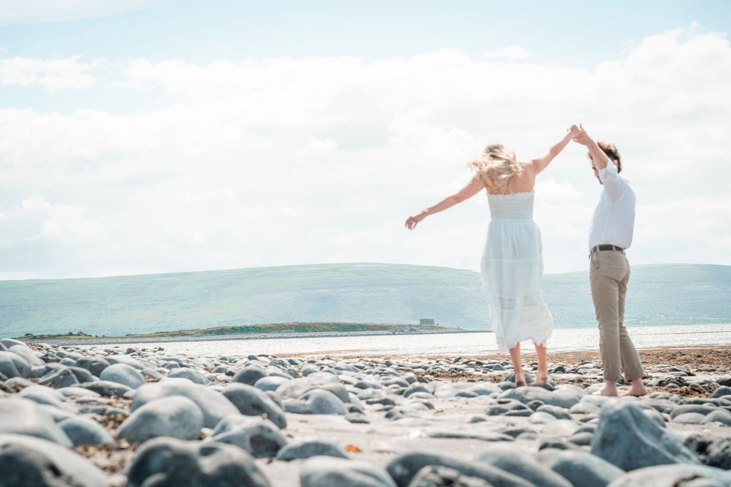 A couple dances together on a rocky beach with mountains visible in the distance.