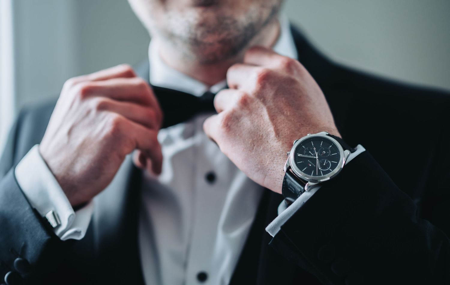 Groom in black tuxedo adjusting bow tie wearing silver chronograph watch on wedding day.