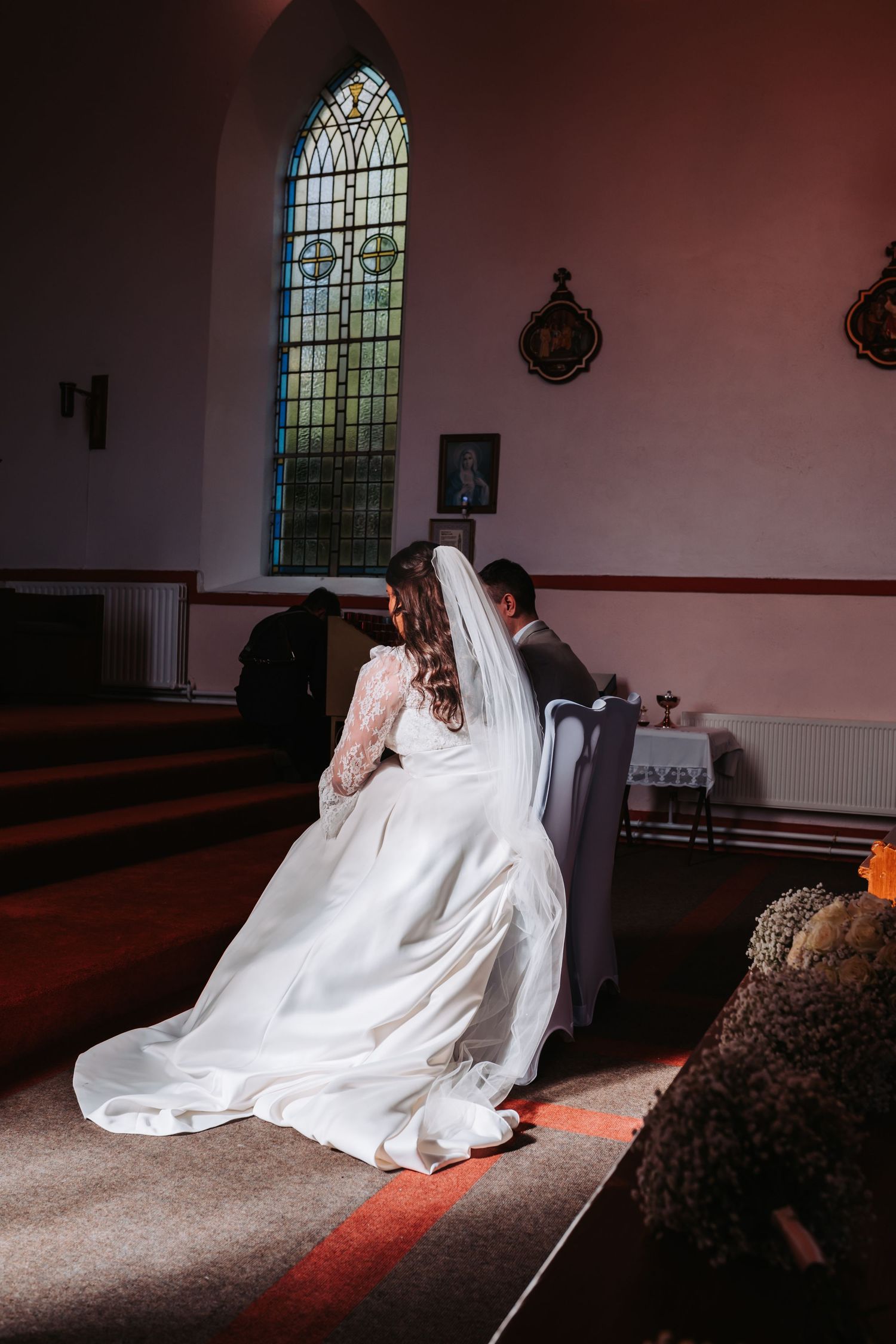 Bride and groom seated together in a sunlit church aisle, bride wearing a flowing white gown and veil near stained glass window.