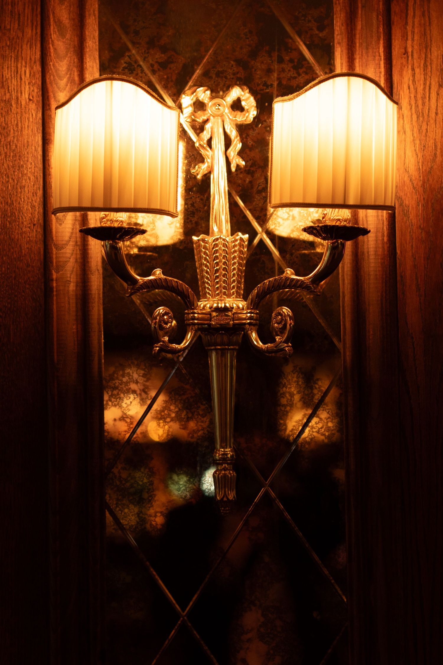 Elegant gold double wall sconce with cream shades and bow detail glowing warmly against mirrored background.