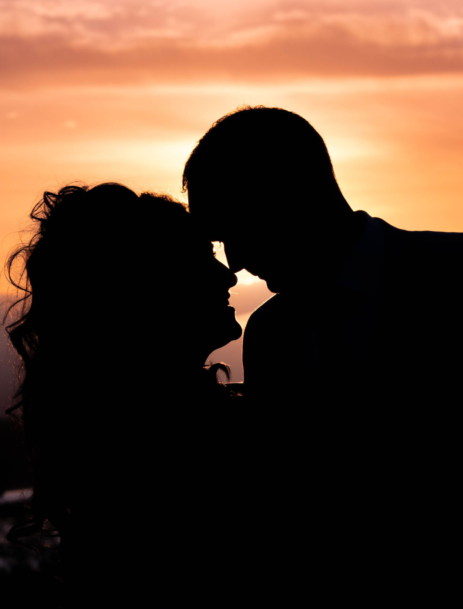Romantic couple silhouette touching foreheads against vibrant orange sunset sky.