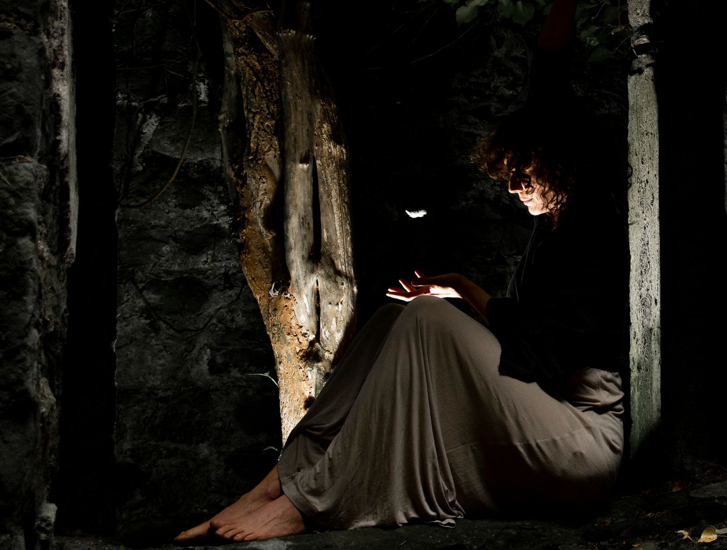 Person reading by moonlight in dark stone ruins, illuminated by soft glow while seated against ancient wall.