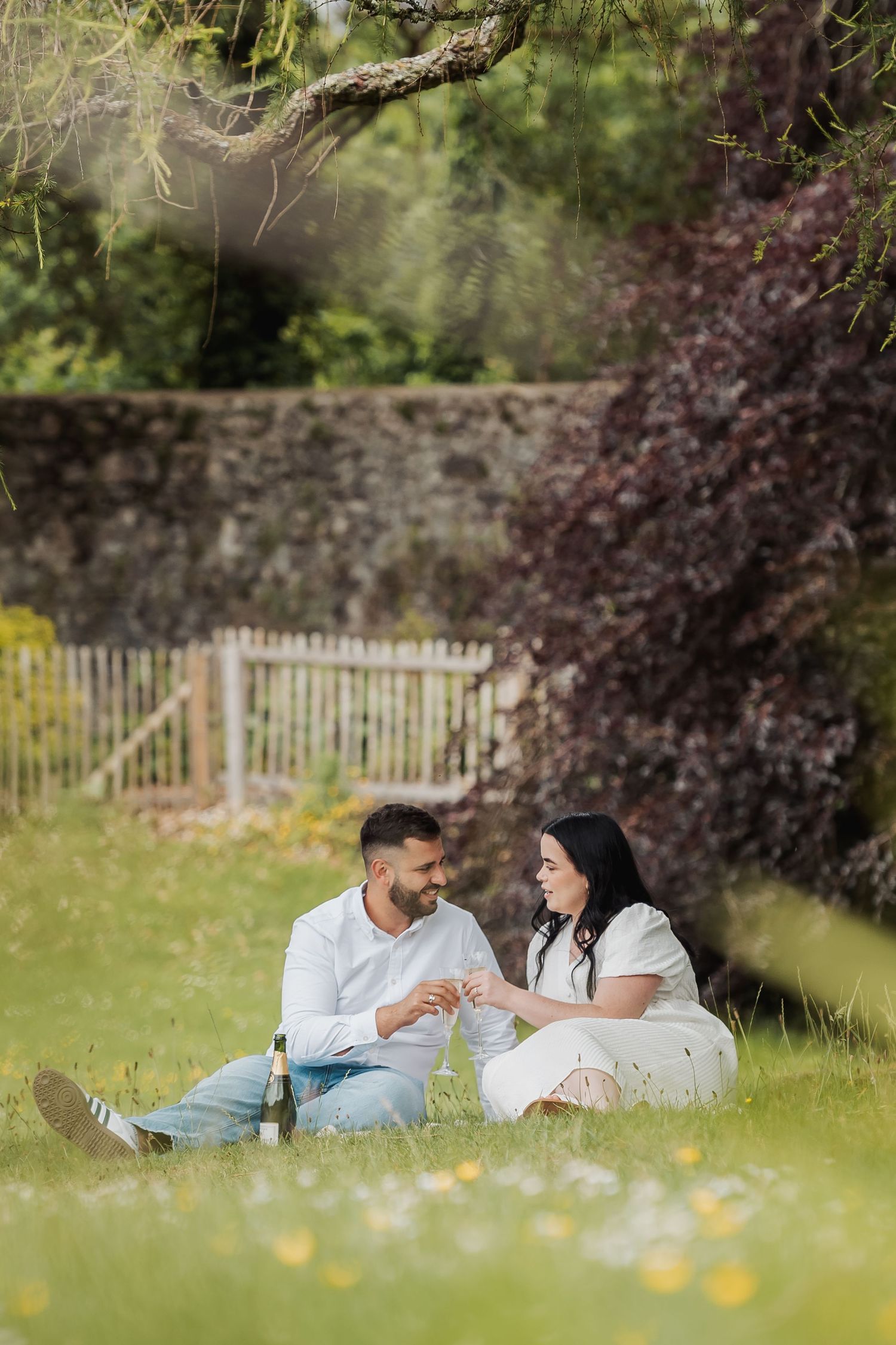 Couple sitting on grass sharing a romantic moment with a bottle of wine in a scenic garden.
