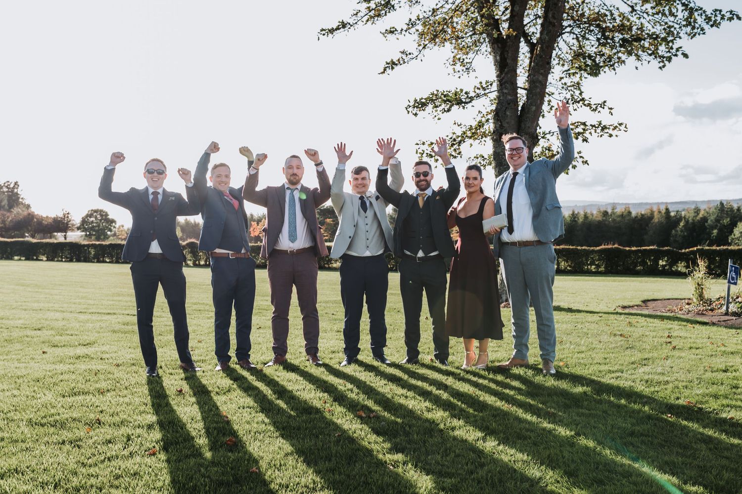 Group of seven people in formal attire celebrating outdoors on a sunny day, raising their arms in joy.