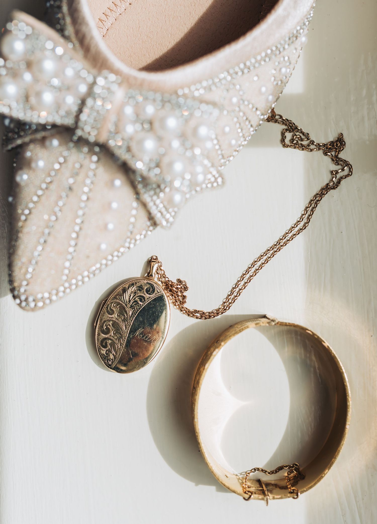 Pearl-adorned bridal shoe with bow, gold locket necklace, and bangle bracelet on white surface.
