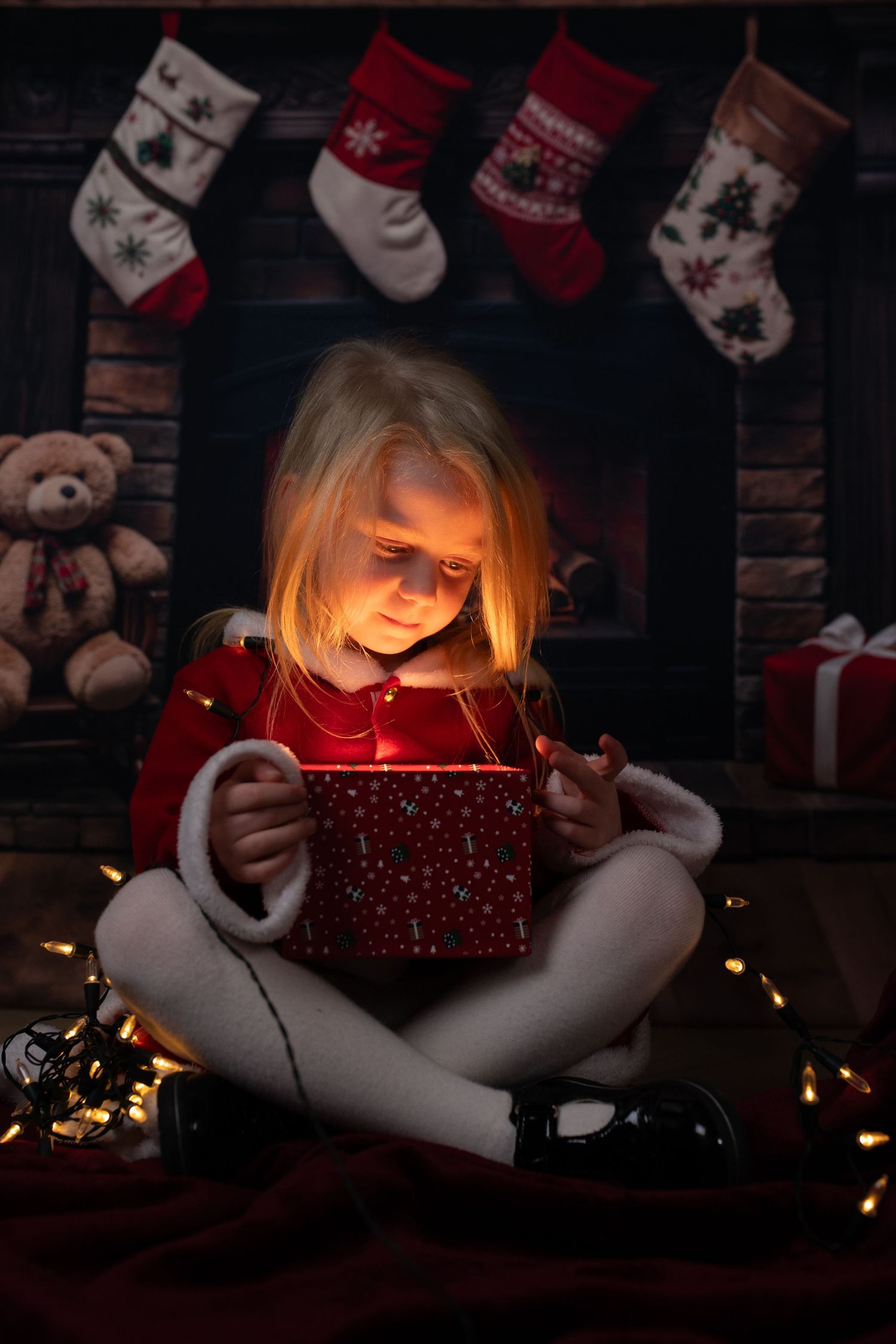 A glowing red Christmas gift box illuminates a dark room with stockings and holiday decorations in the background.