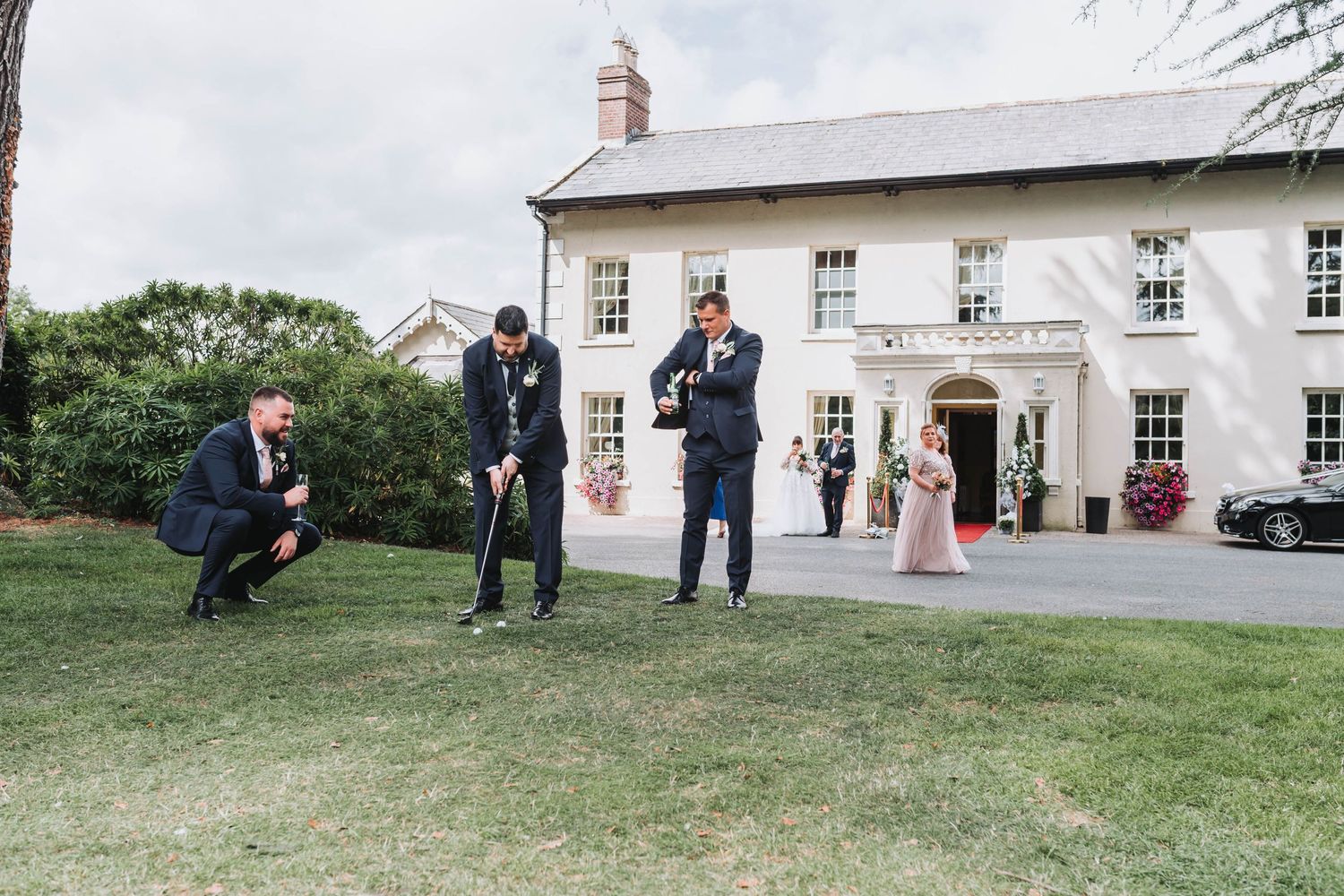 Wedding party members play golf on a grassy lawn outside a white colonial-style building.