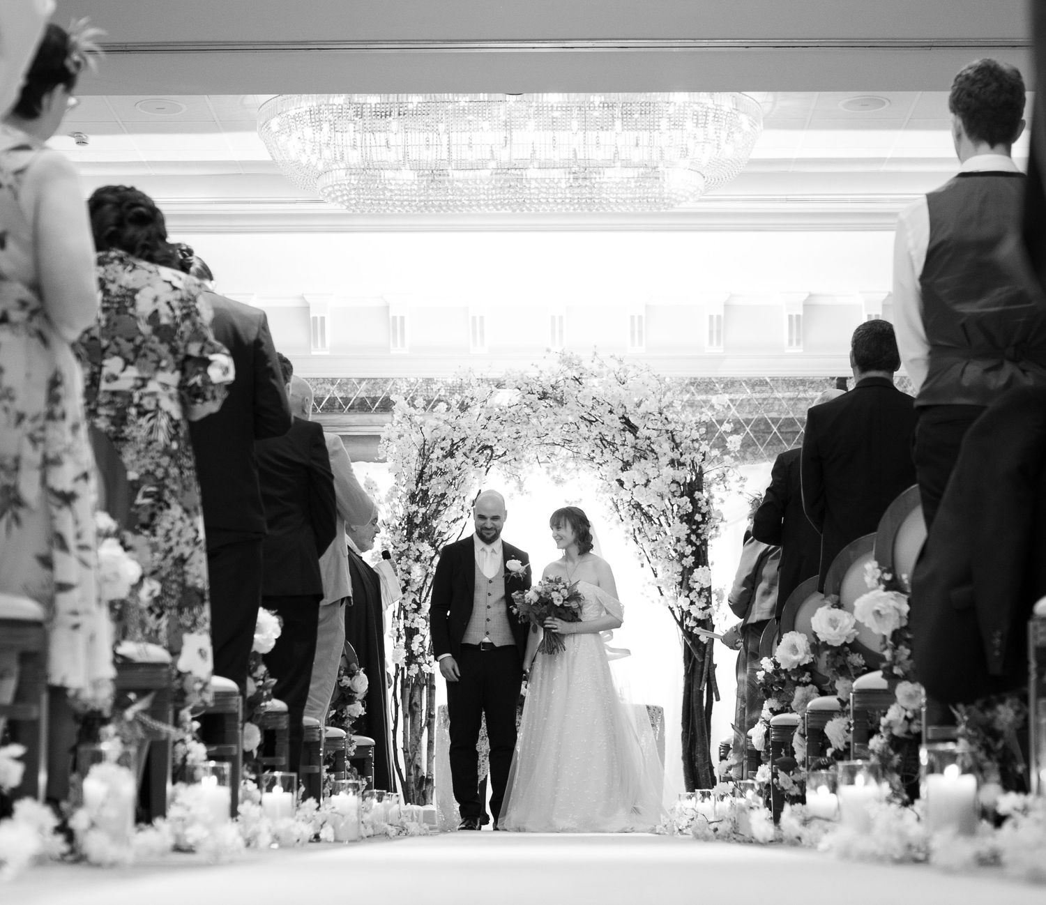 Black and white wedding ceremony photo showing bride and groom at floral arch altar with guests lining the aisle.