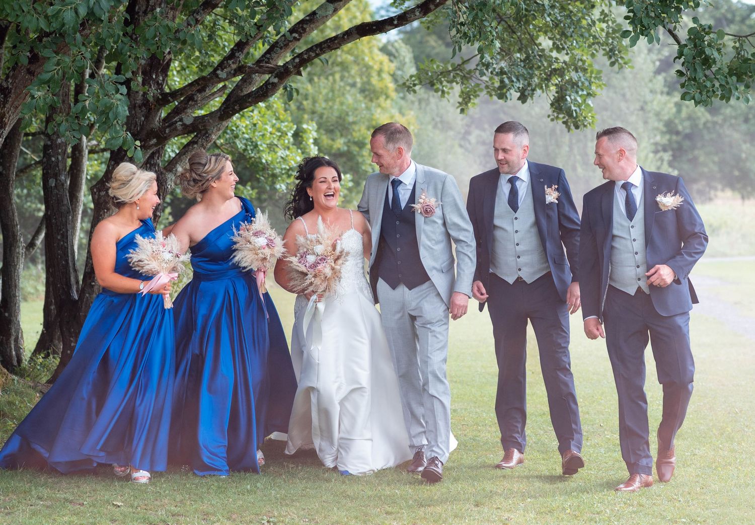 Bride and groom with bridesmaids in blue dresses and groomsmen walking under trees.
