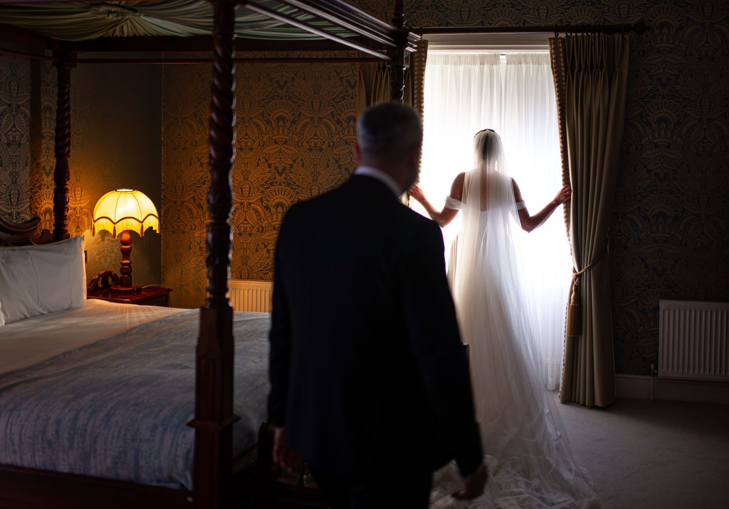 Bride in white wedding gown stands at window in elegant bedroom as groom approaches in dark suit.