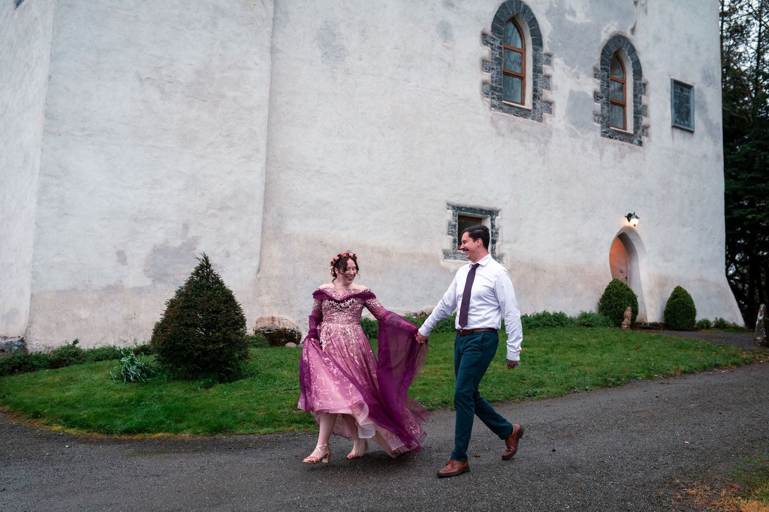 A woman in a purple gown and a man in a suit walk hand in hand outside a white stone building.