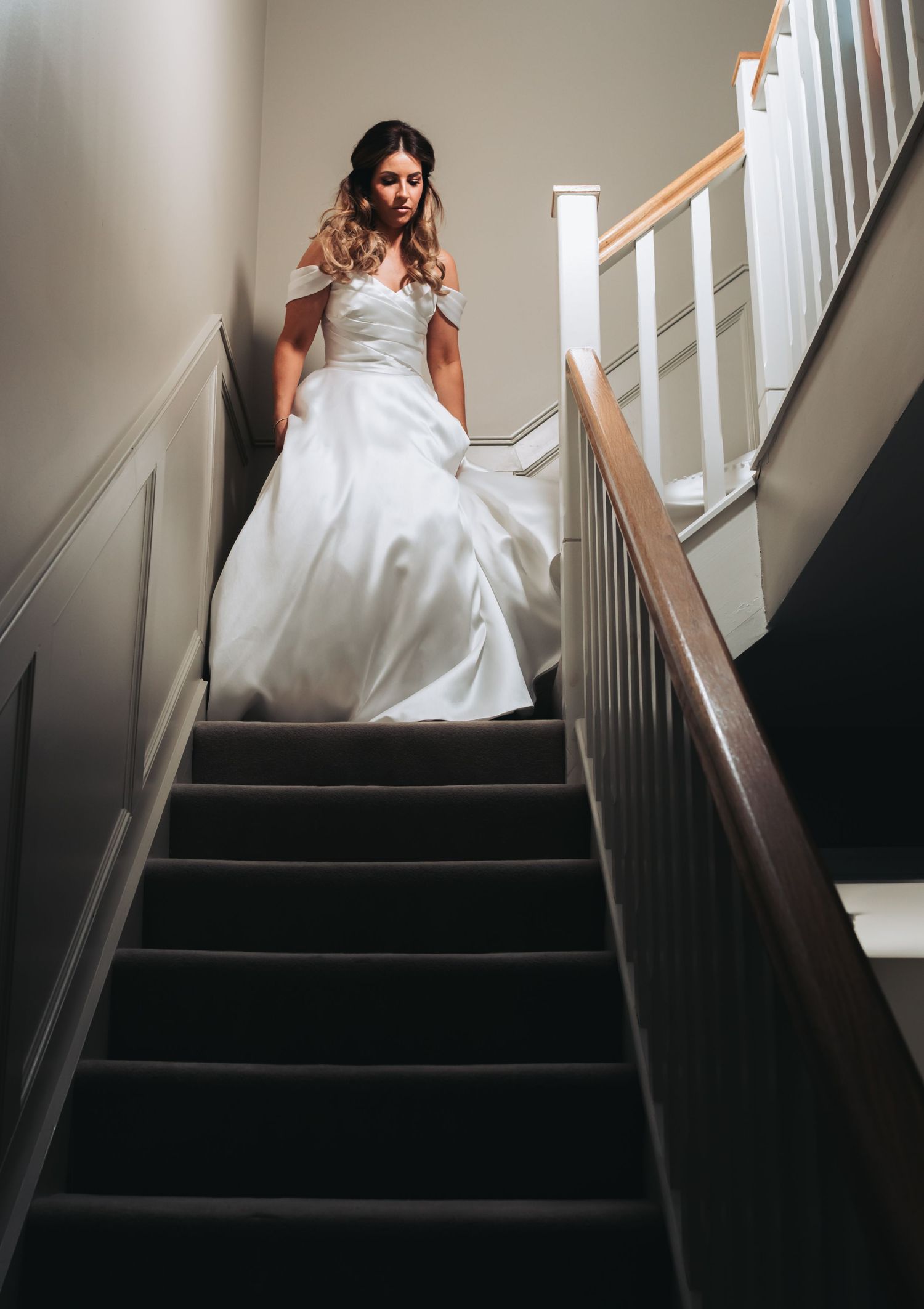Bride in white off-shoulder ball gown descending staircase, looking down elegantly.