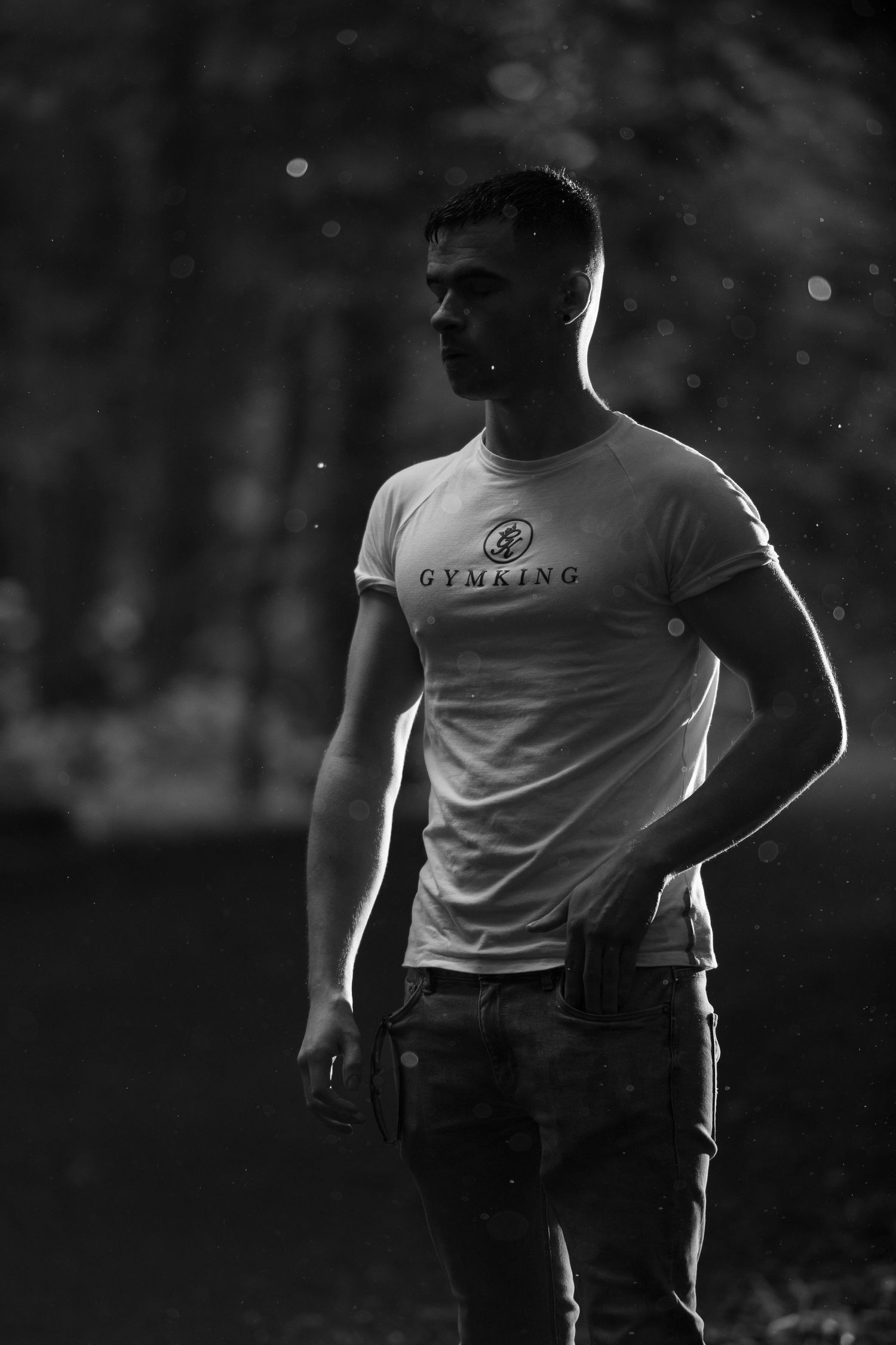 Black and white photo of a muscular man wearing a Gymking t-shirt and jeans, standing outdoors.