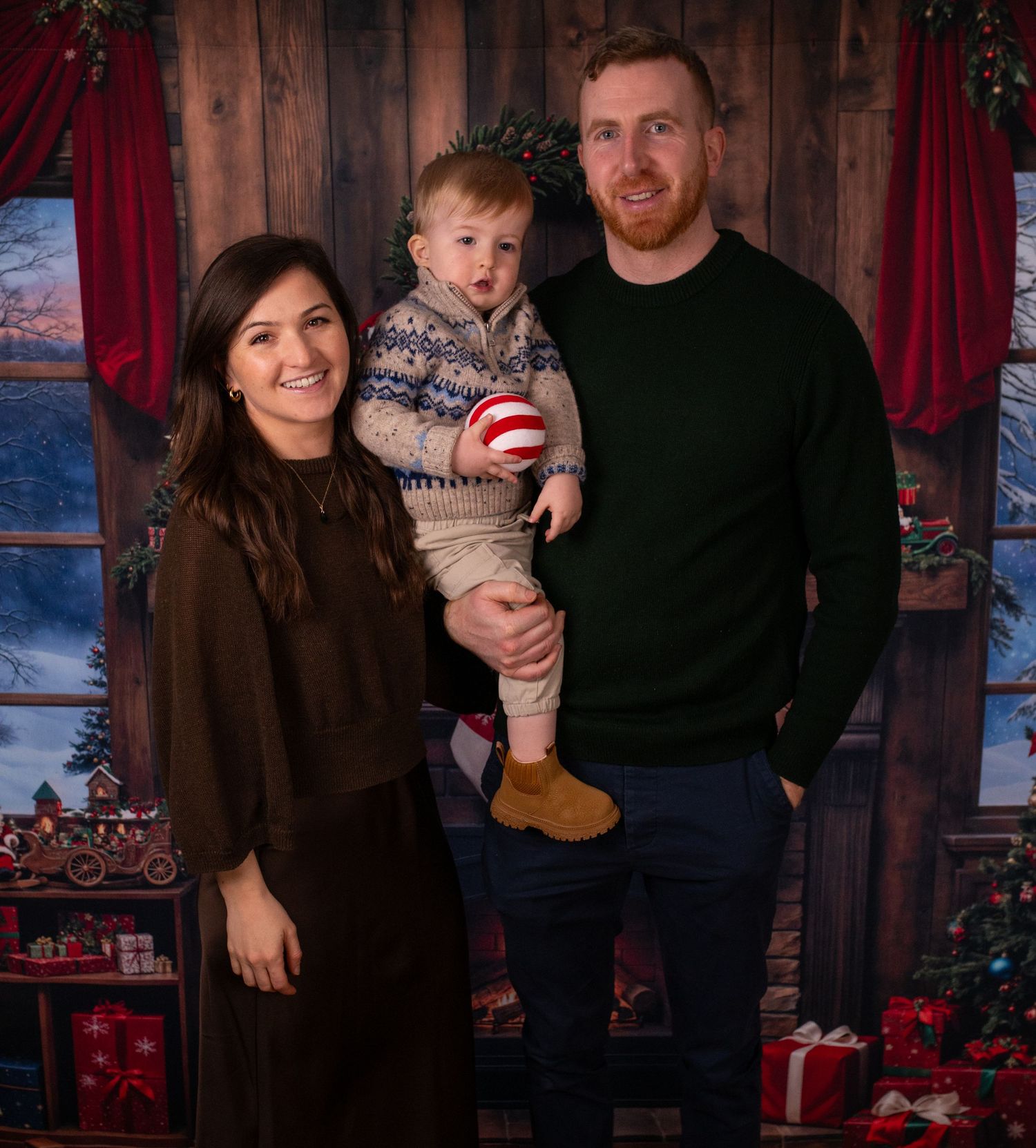 Family poses in front of rustic wooden wall with Christmas decorations and red curtains in background.
