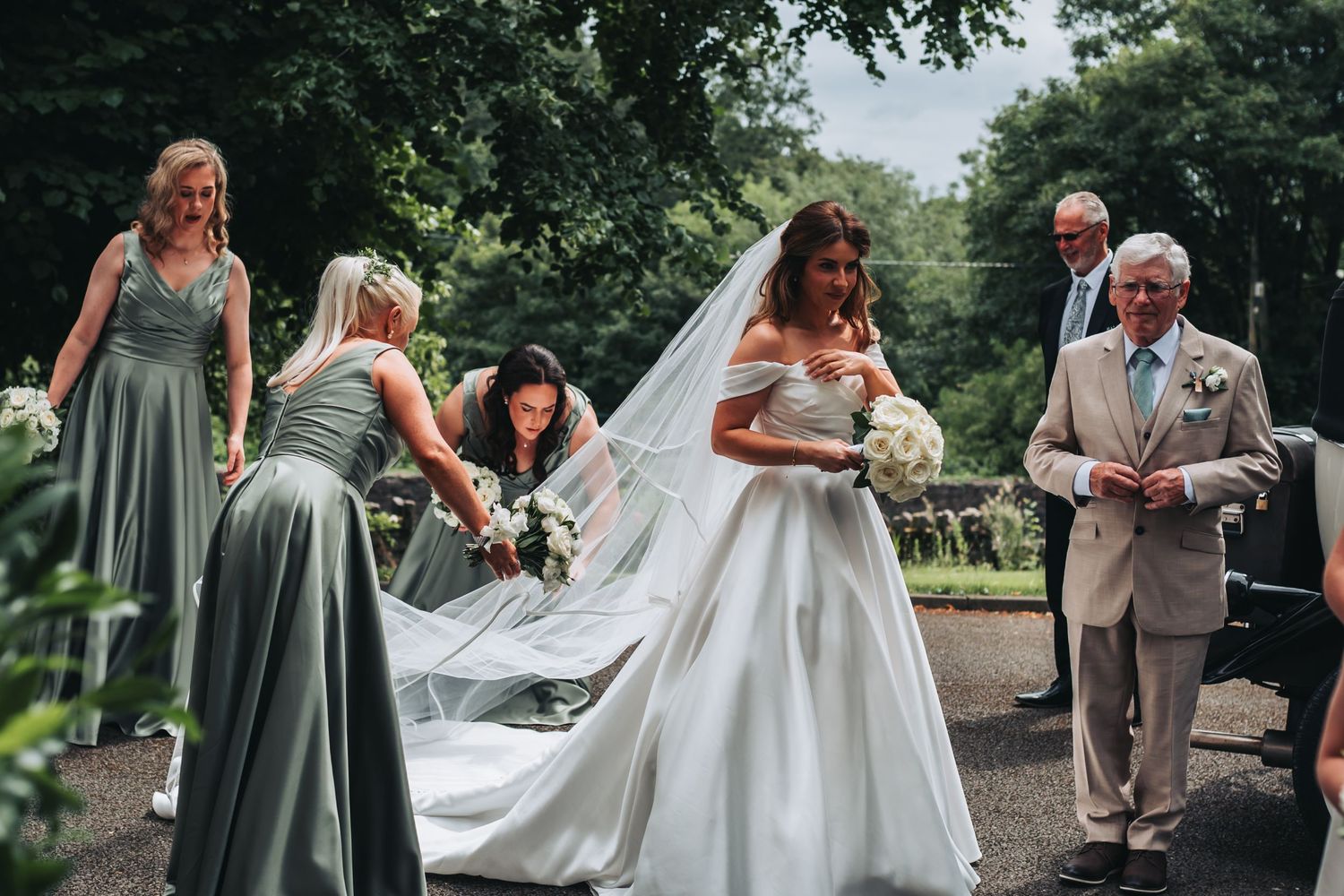 Bridesmaids in sage green dresses adjusting bride's veil outdoors.
