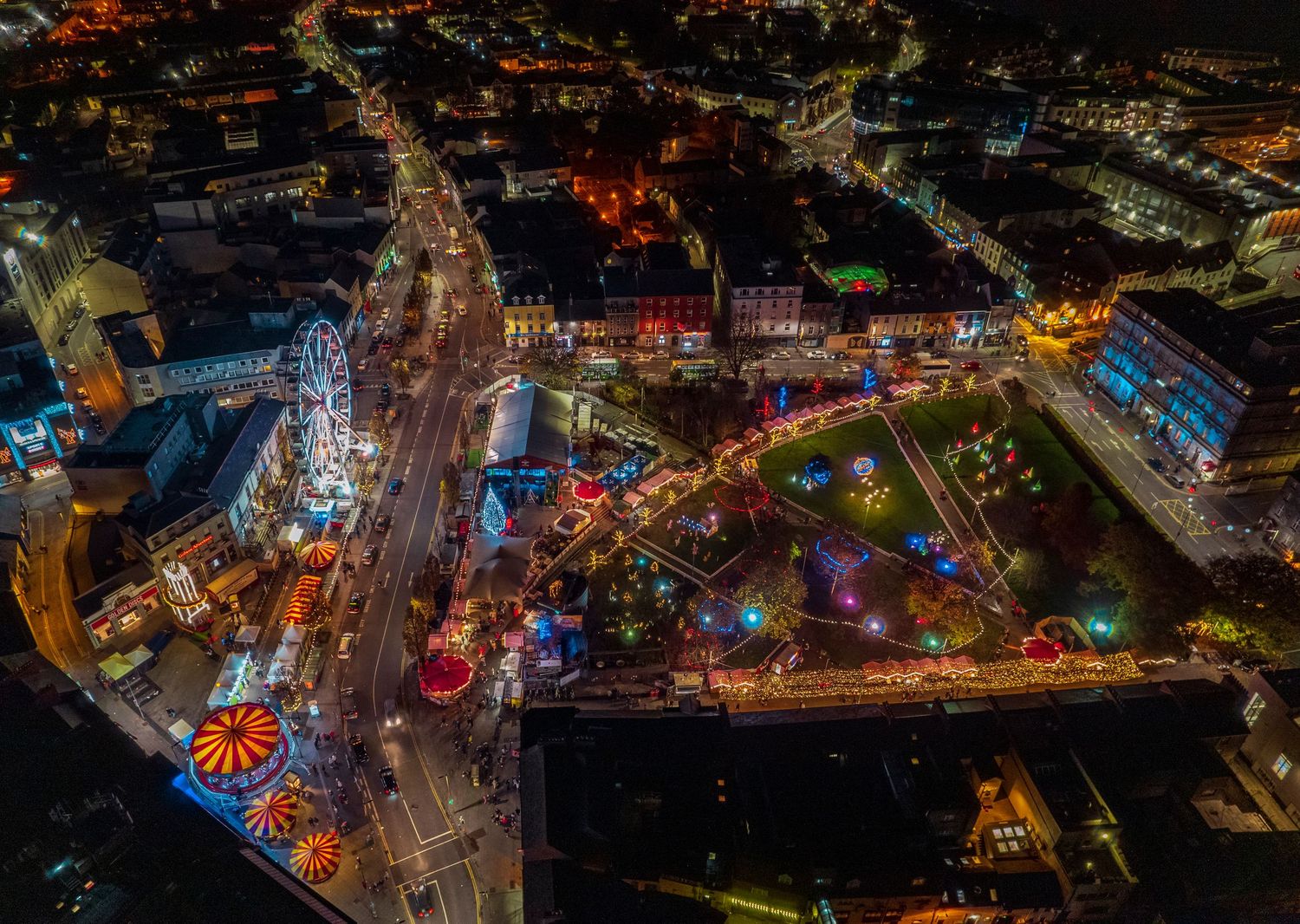 Aerial night view of a festive Christmas market with colorful lights and a Ferris wheel in a busy city center.